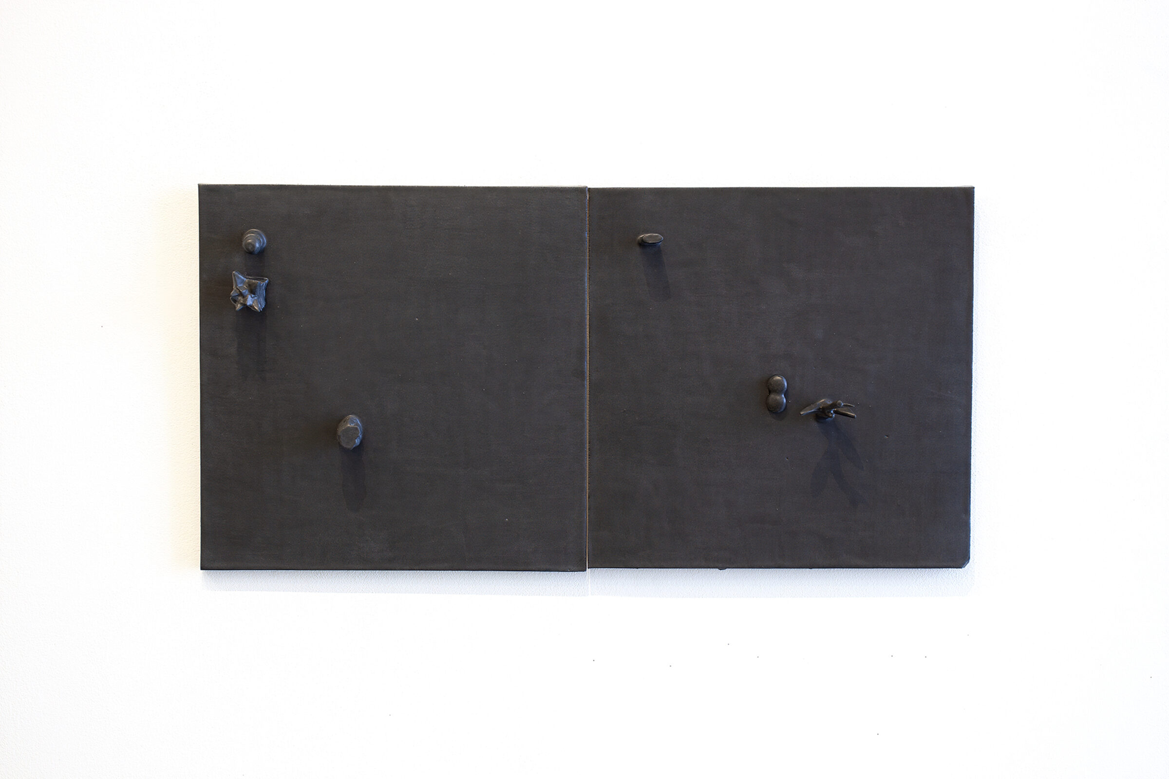 Two dark matte ceramic panels mounted side by side on a white wall, each fitted with metallic knobs and hardware. The left panel features two circular knobs positioned vertically, while the right panel has a similar knob with an additional key or lever mechanism, creating an austere, minimalist composition suggestive of functional cabinetry.