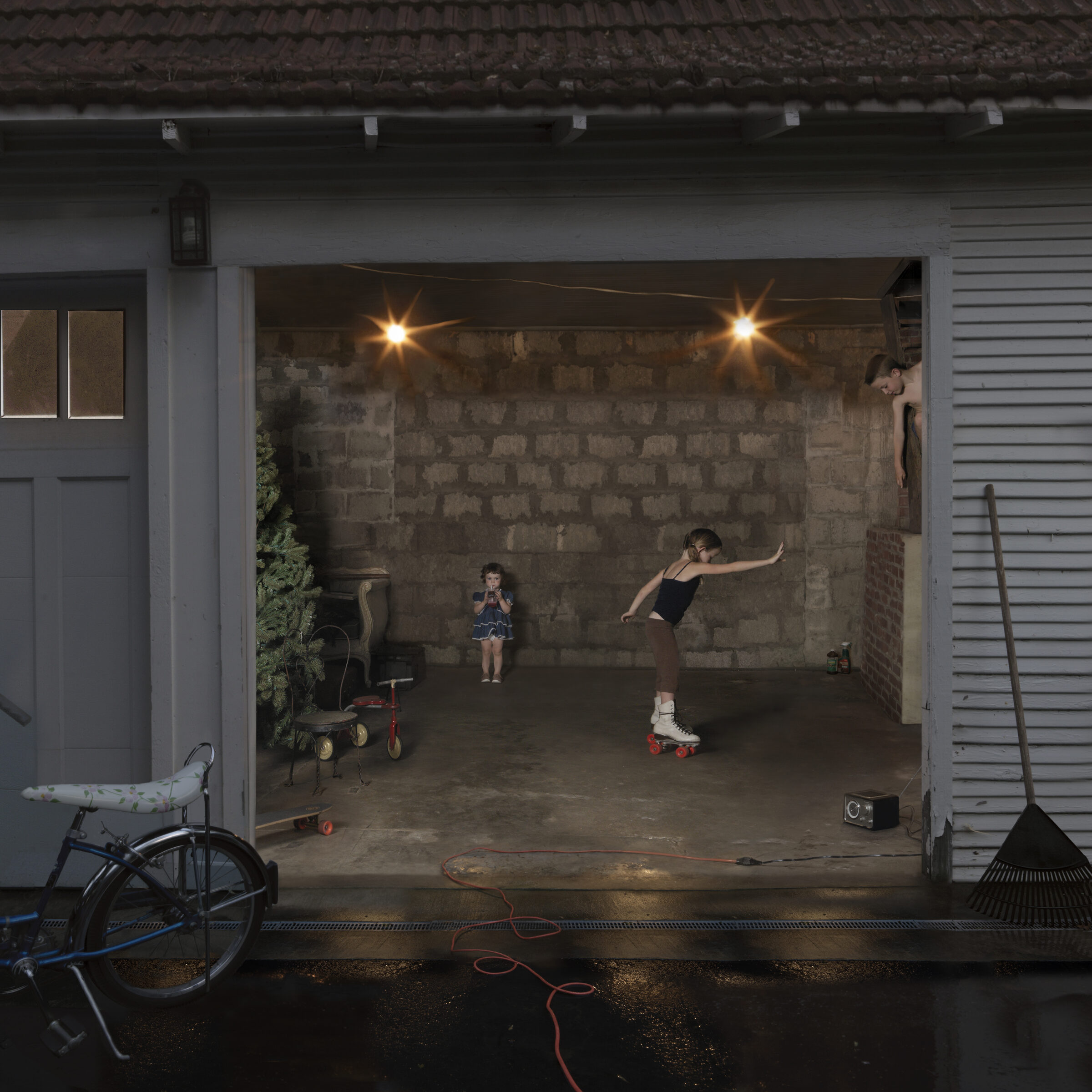 A garage interior photographed at dusk shows two children engaged in play: one wearing roller skates performs a dynamic pose on the concrete floor while another stands nearby in a blue dress. The space is illuminated by two overhead lights, revealing exposed cinder block walls, various toys and household items scattered about including a tricycle and bicycle, and tools leaning against the sides. The photograph captures an intimate domestic moment framed by the open garage door.