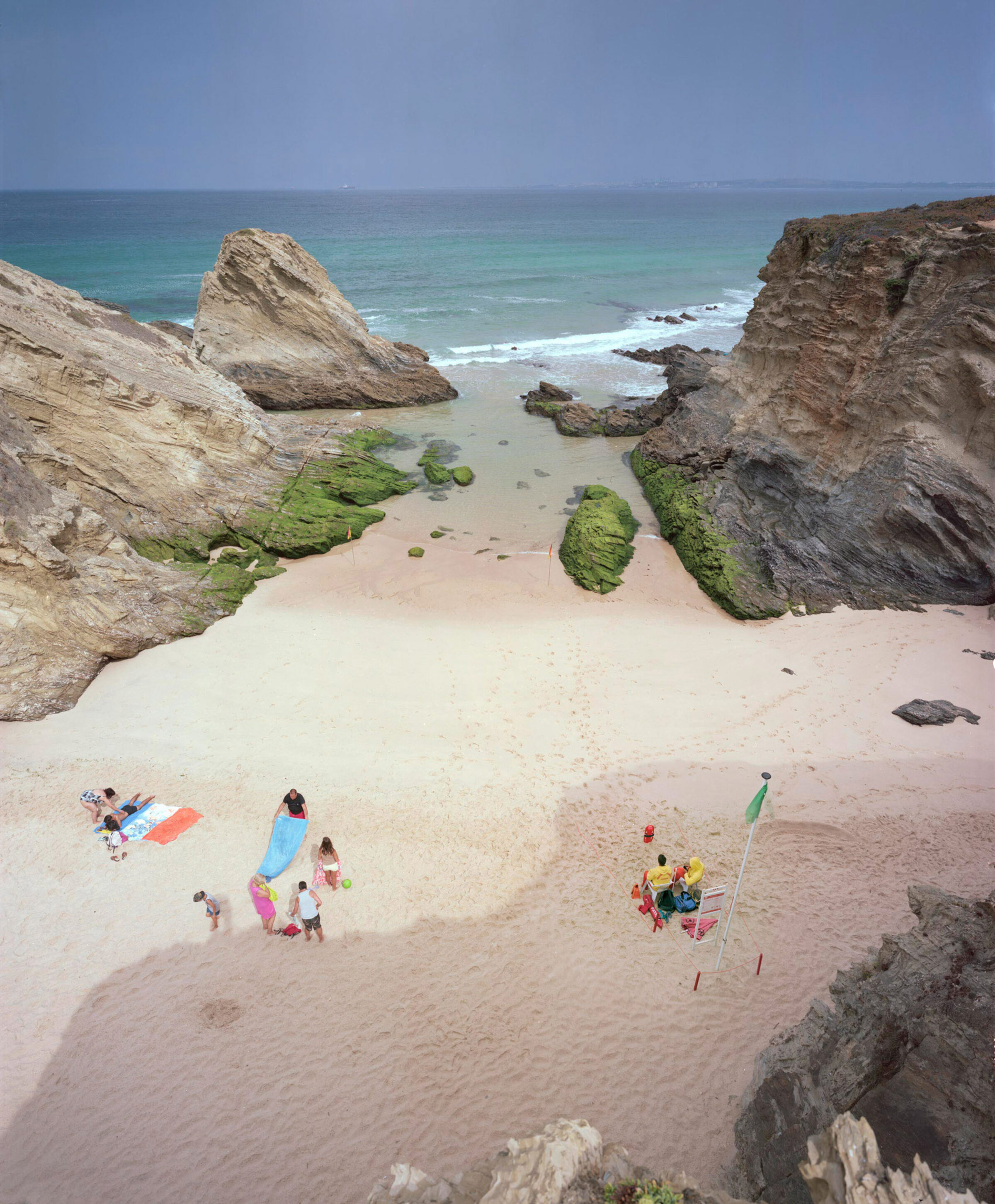 Aerial view of a secluded beach cove framed by dramatic layered rock formations, with turquoise water and white sand. Small groups of beachgoers are scattered across the shore, some with colorful towels and beach equipment, while moss-covered rocks and boulders dot the sandy beach and shallow waters.