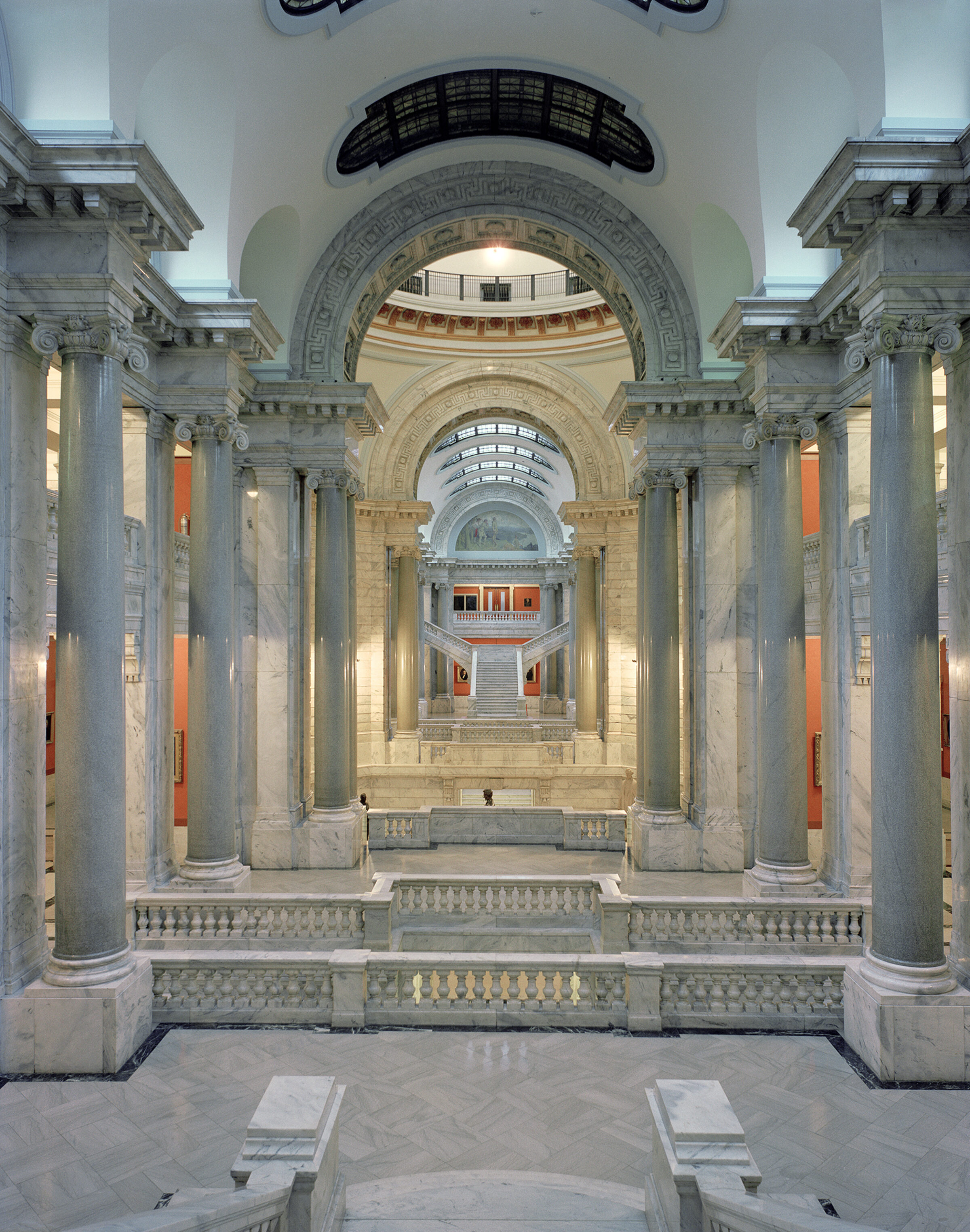 A symmetrical corridor with towering fluted columns in pale marble flanks a central passage, leading to a grand arched rotunda with ornamental detailing. Warm lighting illuminates the neoclassical architecture, while burnt orange accent panels and ornate balustrades recede into the distance.