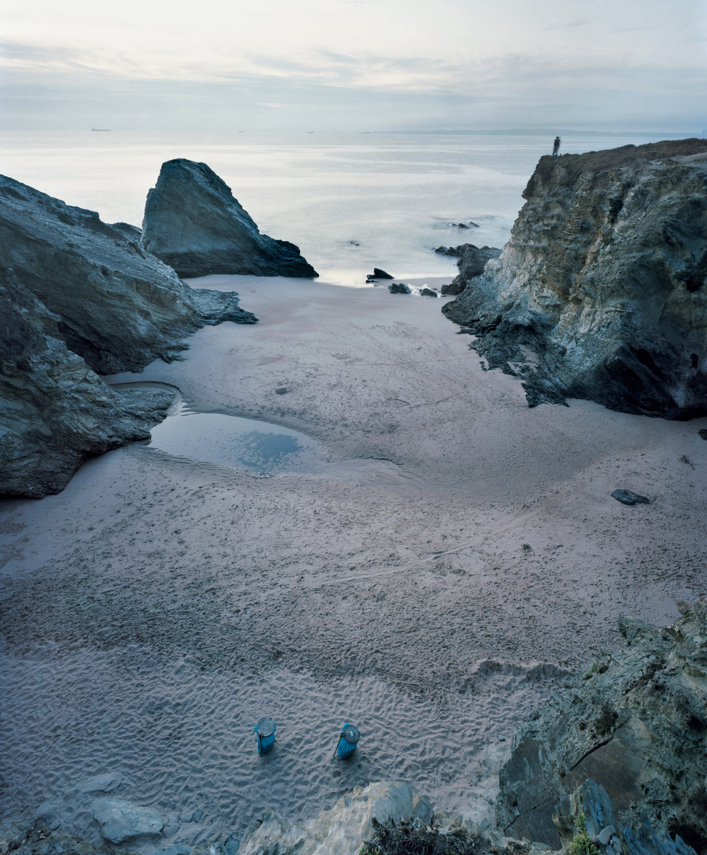 A coastal landscape at dusk featuring a sandy beach bordered by dramatic grey rock formations and sea stacks. Two small blue objects sit on the wet sand in the foreground, while a solitary figure stands on a distant cliff to the right. The calm, pale waters and overcast sky create a cool, muted color palette of greys, blues, and whites.