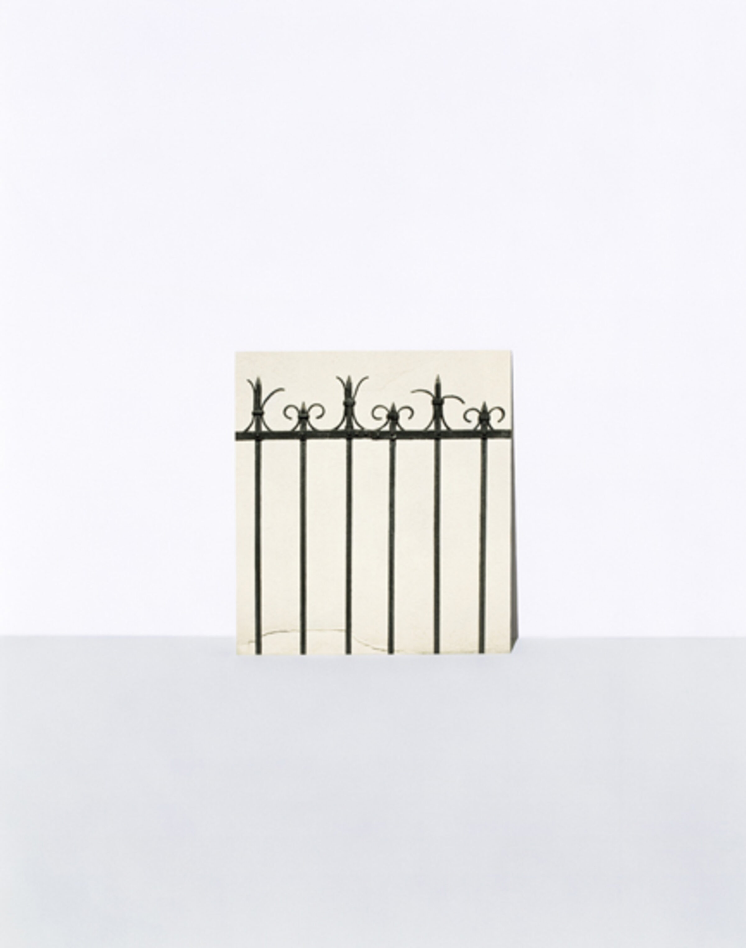 A minimalist composition featuring a decorative wrought iron fence with ornamental finials positioned against a pale cream-colored wall, casting sharp shadows on a light gray ground plane below.