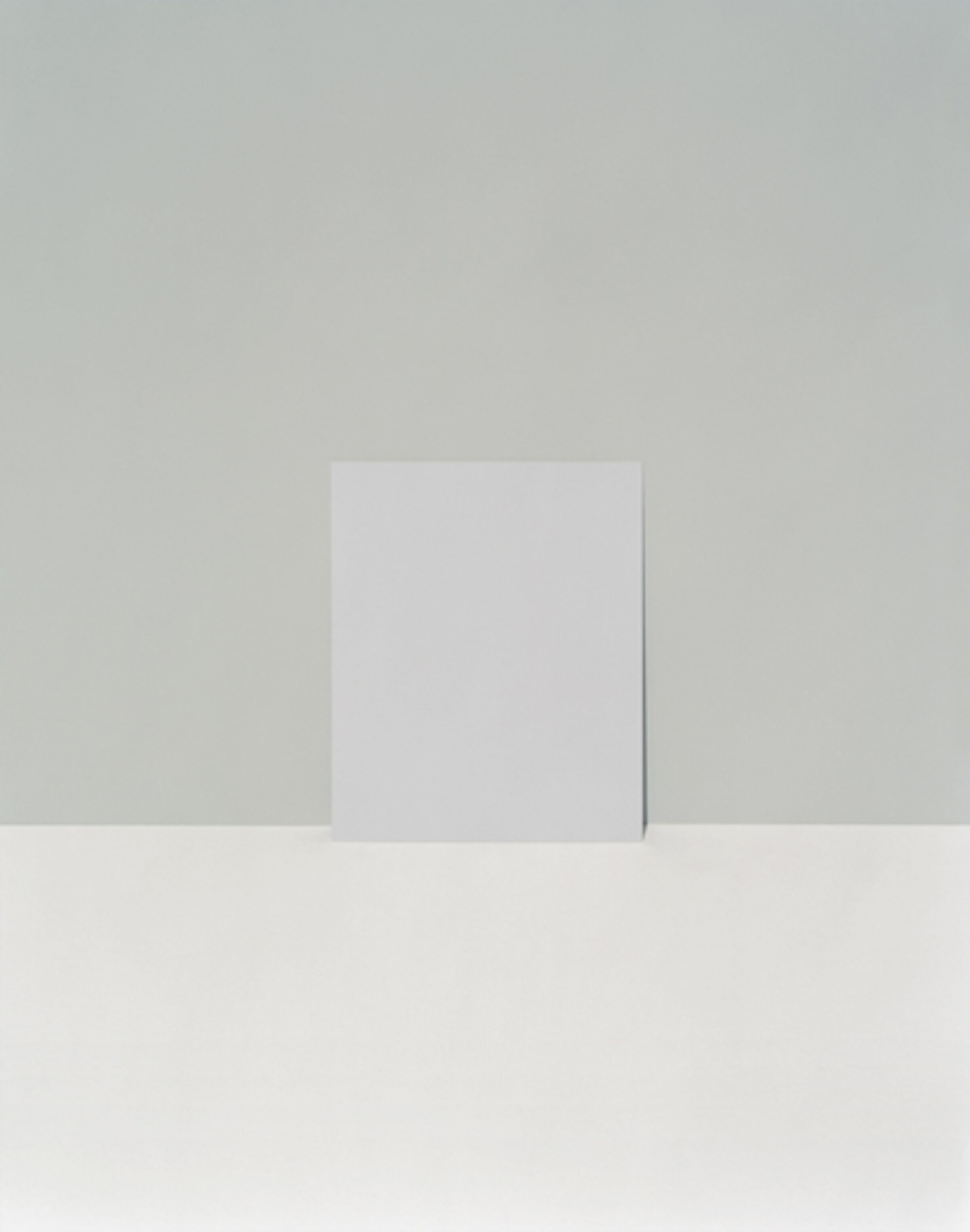 A minimalist composition featuring a white rectangular form standing vertically against a soft gray wall, casting a subtle shadow. The work employs a restrained palette of grays and whites with precise geometric forms, emphasizing negative space and the quiet interplay of light and shadow.