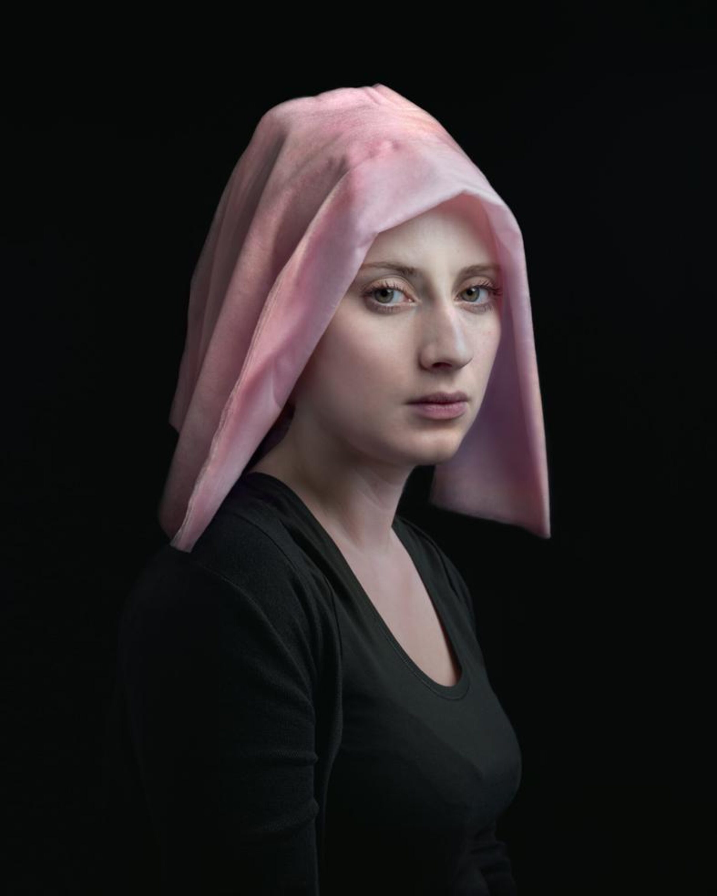 Portrait of a young woman in profile against a black background, wearing a black long-sleeved top and a draped pink fabric covering her head. Her pale complexion and direct gaze create a contemplative mood, with soft lighting emphasizing the delicate folds of the translucent cloth.