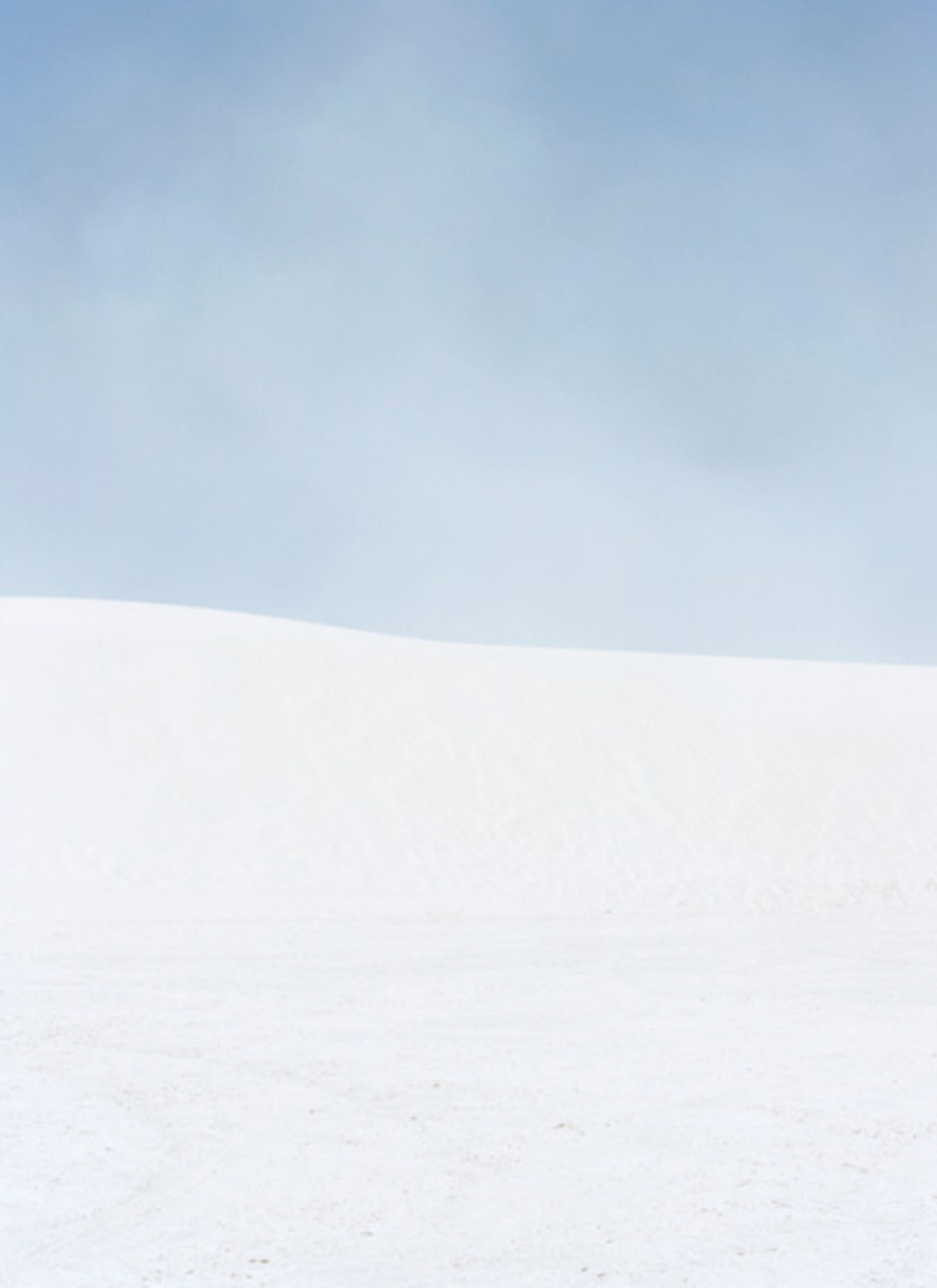 A minimalist landscape divided horizontally into two zones: a soft blue-gray sky occupying the upper portion, and a vast expanse of white snow or sand below, with subtle variations in tone creating gentle undulations across the lower plane.