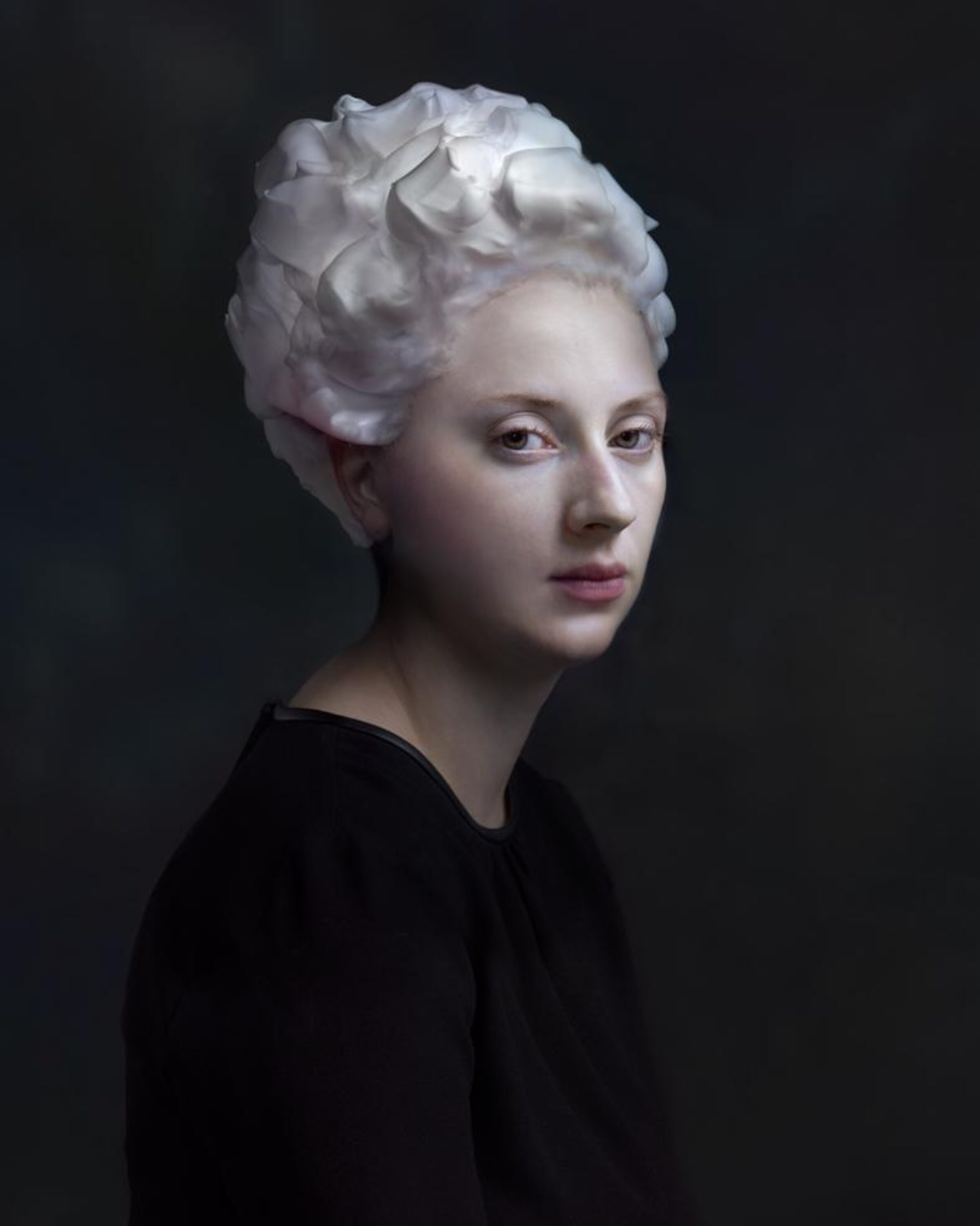 Portrait of a person with pale skin and white sculptural hair styled in voluminous waves, wearing a black garment against a dark background. The lighting is soft and dramatic, emphasizing the figure's serene expression and the luminous quality of the hair.