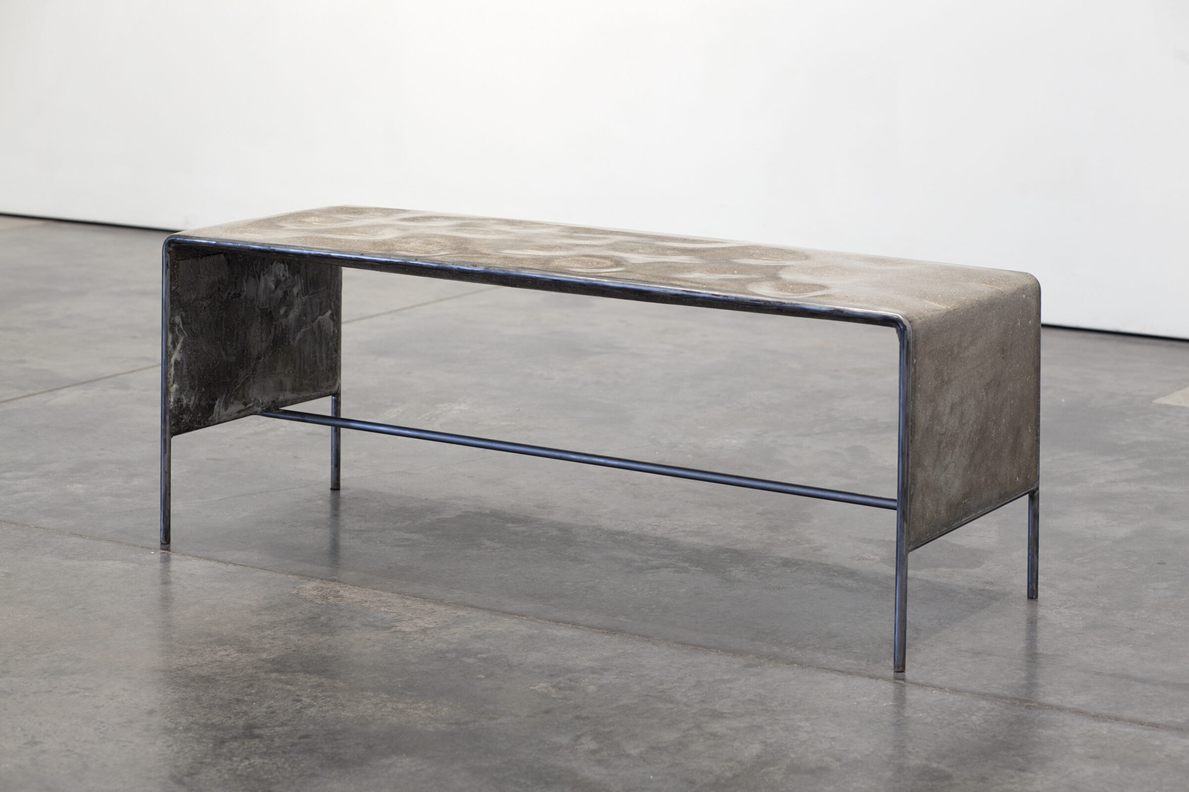 Industrial bench with a concrete and ceramic top featuring geometric surface patterns, supported by dark metal legs and frame with blue-tinted steel accents. The weathered materials and minimalist construction create a stark, contemporary sculptural form displayed on polished concrete flooring.