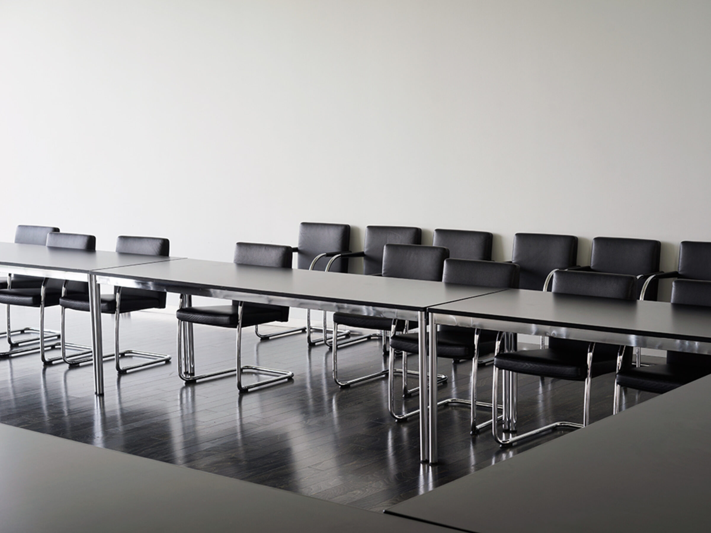A minimalist interior photograph depicting a sleek conference room with a long, reflective dark table surrounded by black leather chairs with chrome frames. The polished dark flooring mirrors the table's surface, while white walls and subtle overhead lighting create a stark, contemporary aesthetic emphasizing geometric forms and material contrasts.