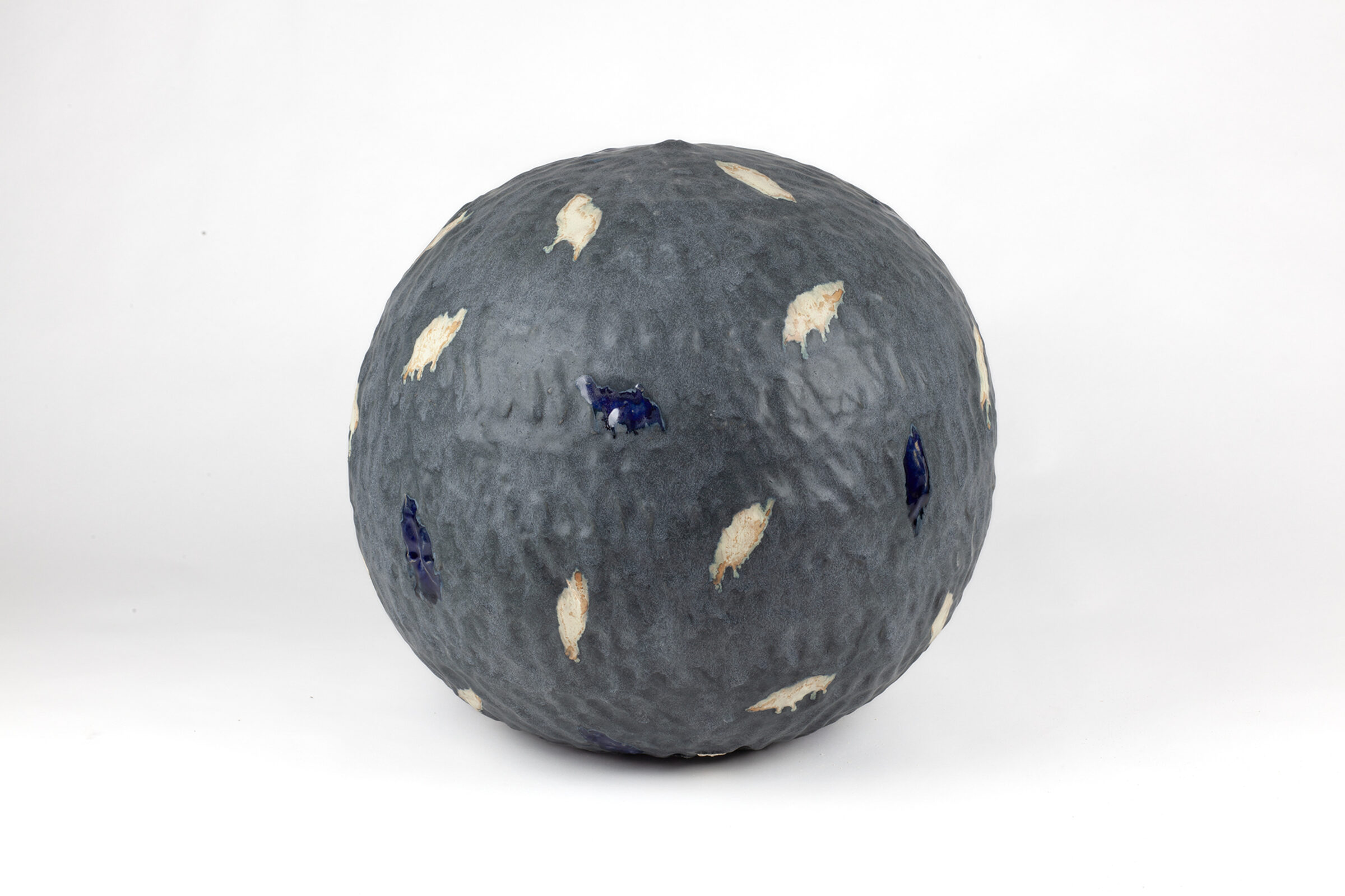 A spherical ceramic form with a textured grey surface scattered with cream-colored and deep blue leaf-shaped appliqués, displayed against a white background.