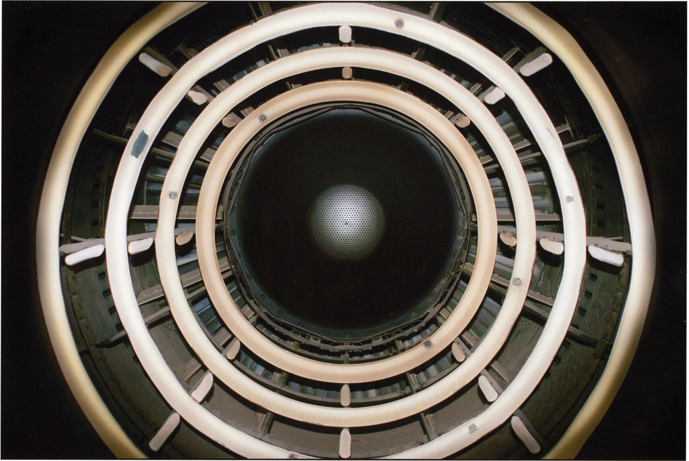 Concentric circles of cream and white rings with metal fixtures frame a dark central aperture, creating a geometric spiral effect. The composition suggests looking down into a cylindrical industrial or architectural structure, with alternating bands of light and shadow receding toward a barely visible illuminated center.