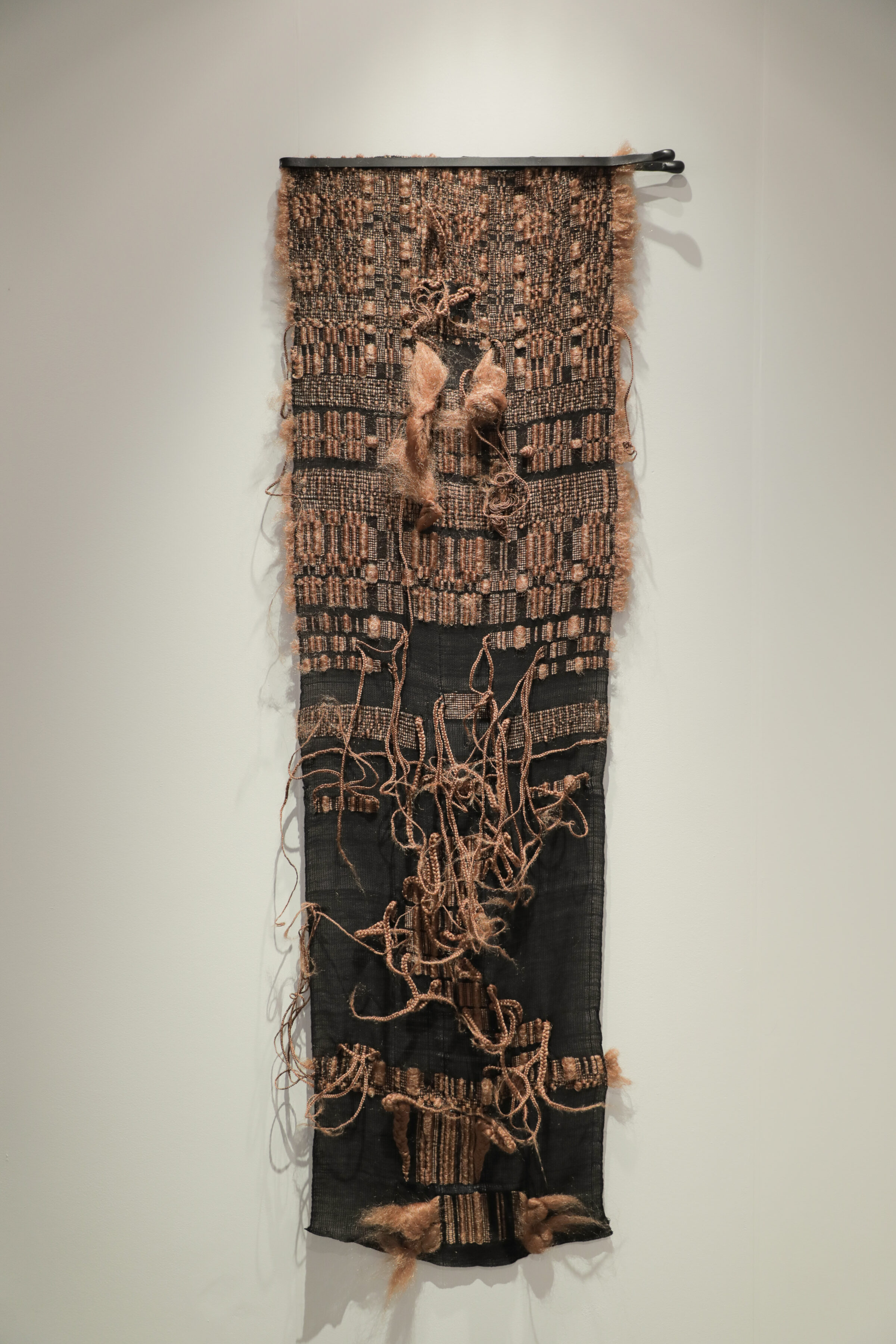 A vertical textile wall hanging combining woven cotton with synthetic hair elements. The upper portion features a tan and black woven pattern with figurative forms, while the lower section is predominantly black with looping strands of blonde hair creating organic, gestural marks across the surface.