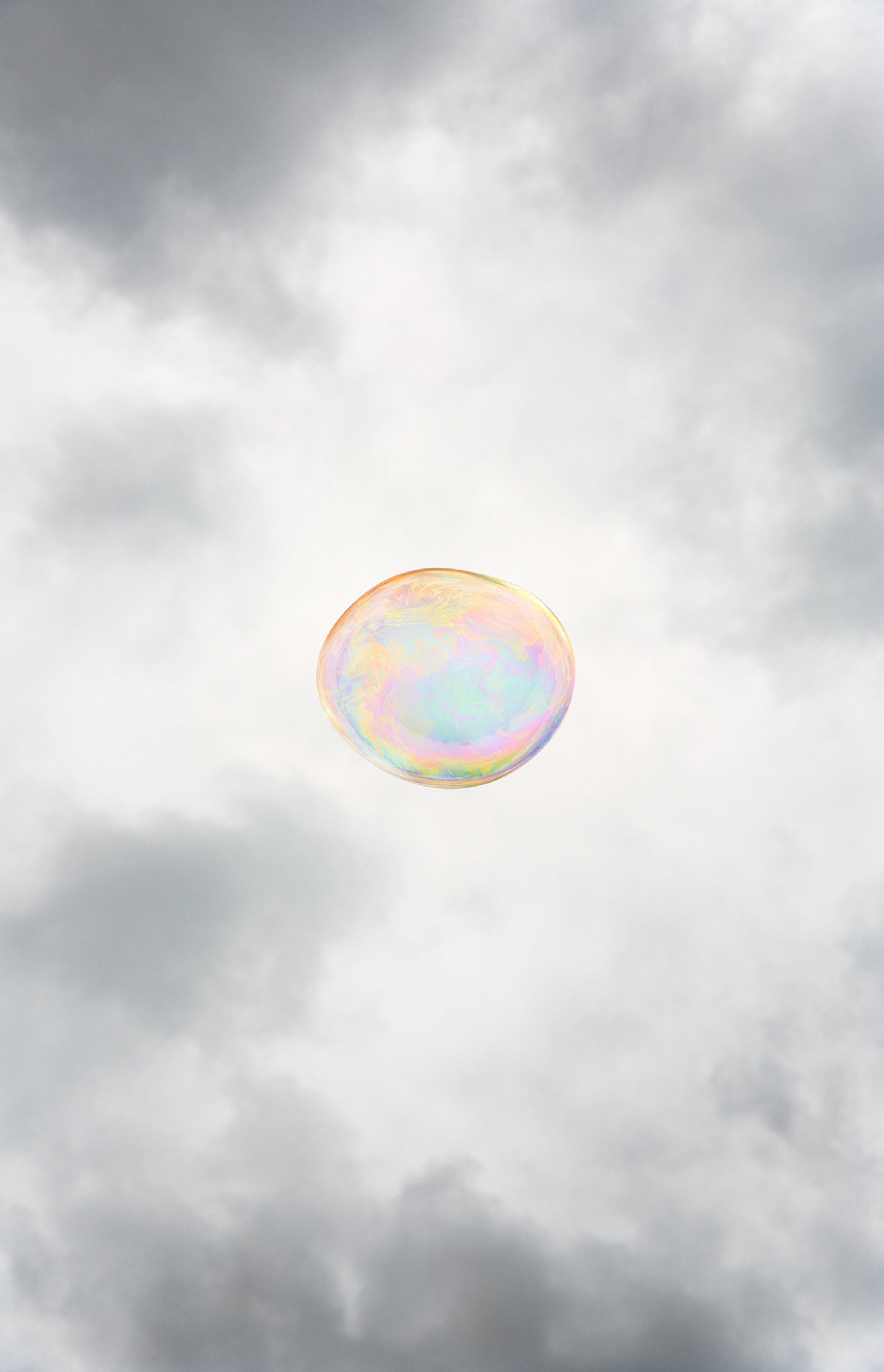 A single iridescent soap bubble floats against a cloudy gray sky, its translucent surface displaying soft pastel hues of pink, yellow, and blue that shimmer across its spherical form.