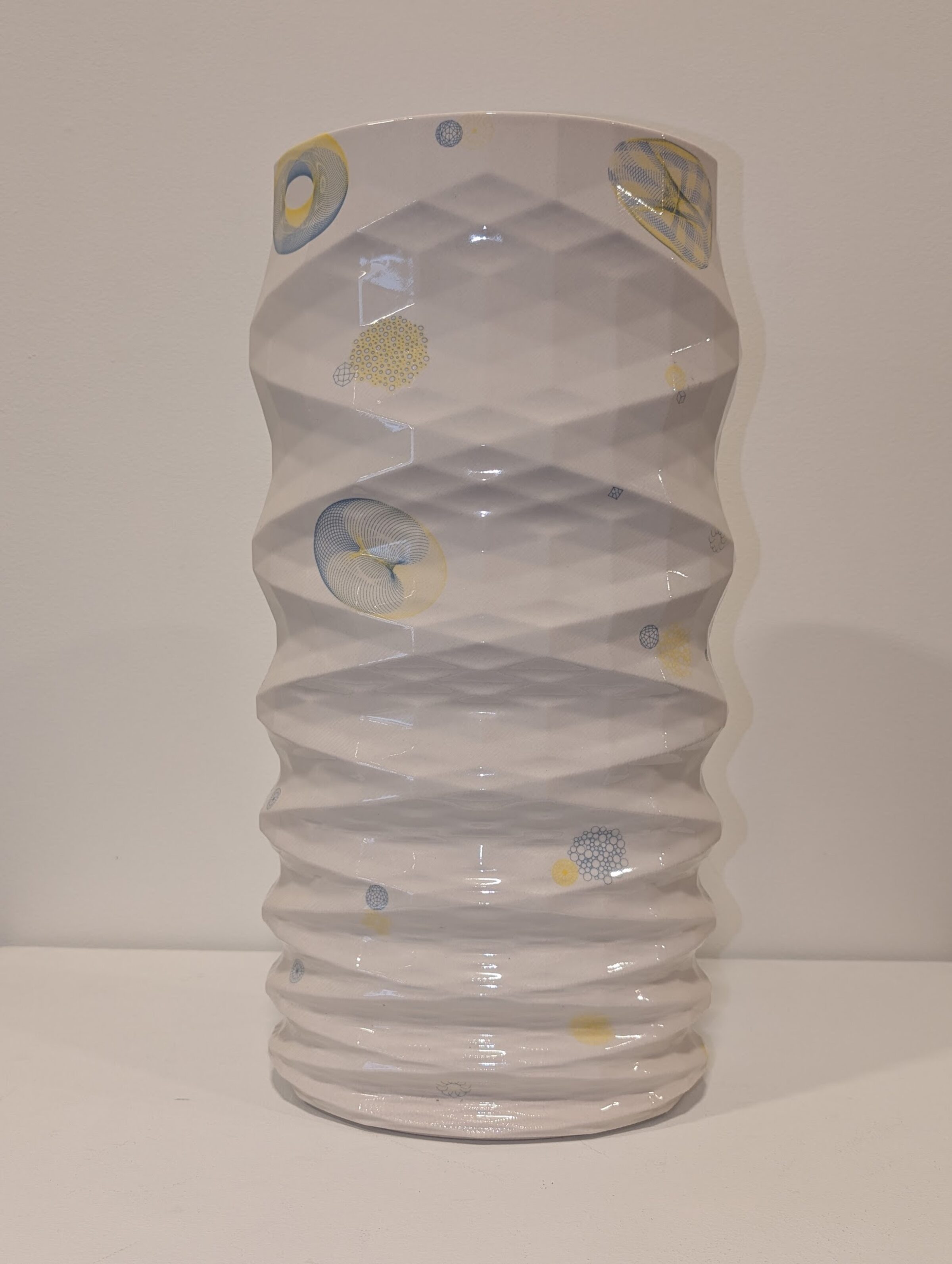 A white porcelain vessel with undulating, organic sides features abstract motifs in soft blue and pale yellow throughout—circular forms, linear patterns, and gestural marks that evoke scientific or natural imagery. The glazed surface catches light across its textured, rhythmically rippled form.