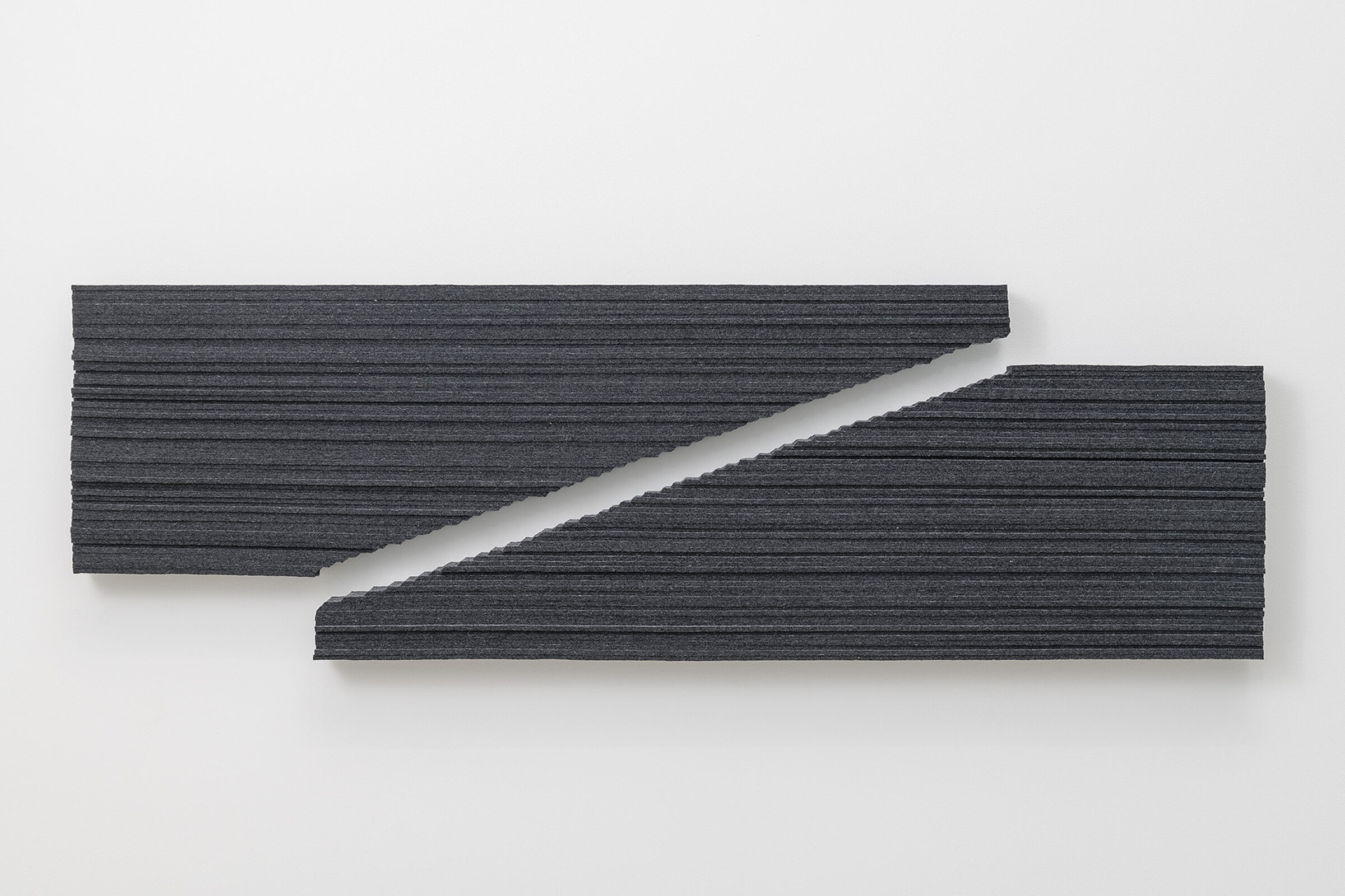 Two dark gray textile strips with horizontal ribbed texture are mounted on a white wall, angled diagonally across one another to create an interlocking geometric composition with a white gap between them.