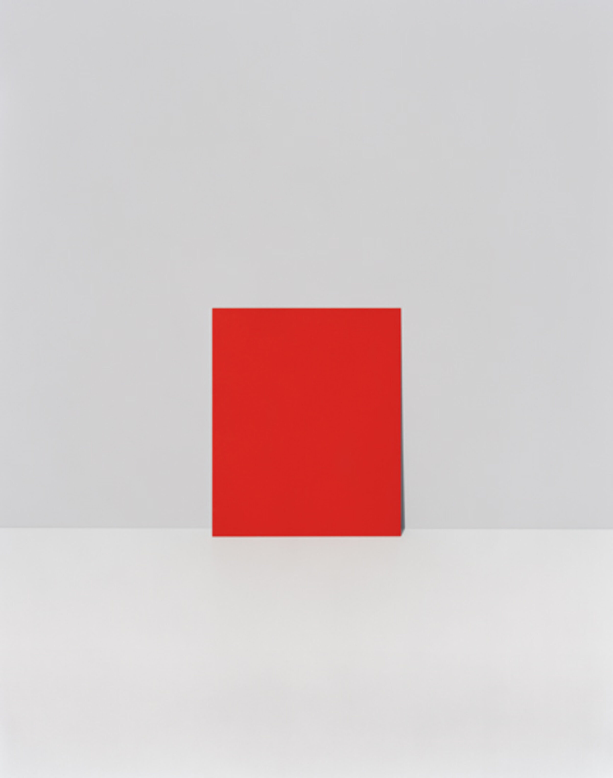 A bright red square positioned centrally against a neutral gray and white background, creating a stark geometric composition with minimal color variation and clean spatial division.