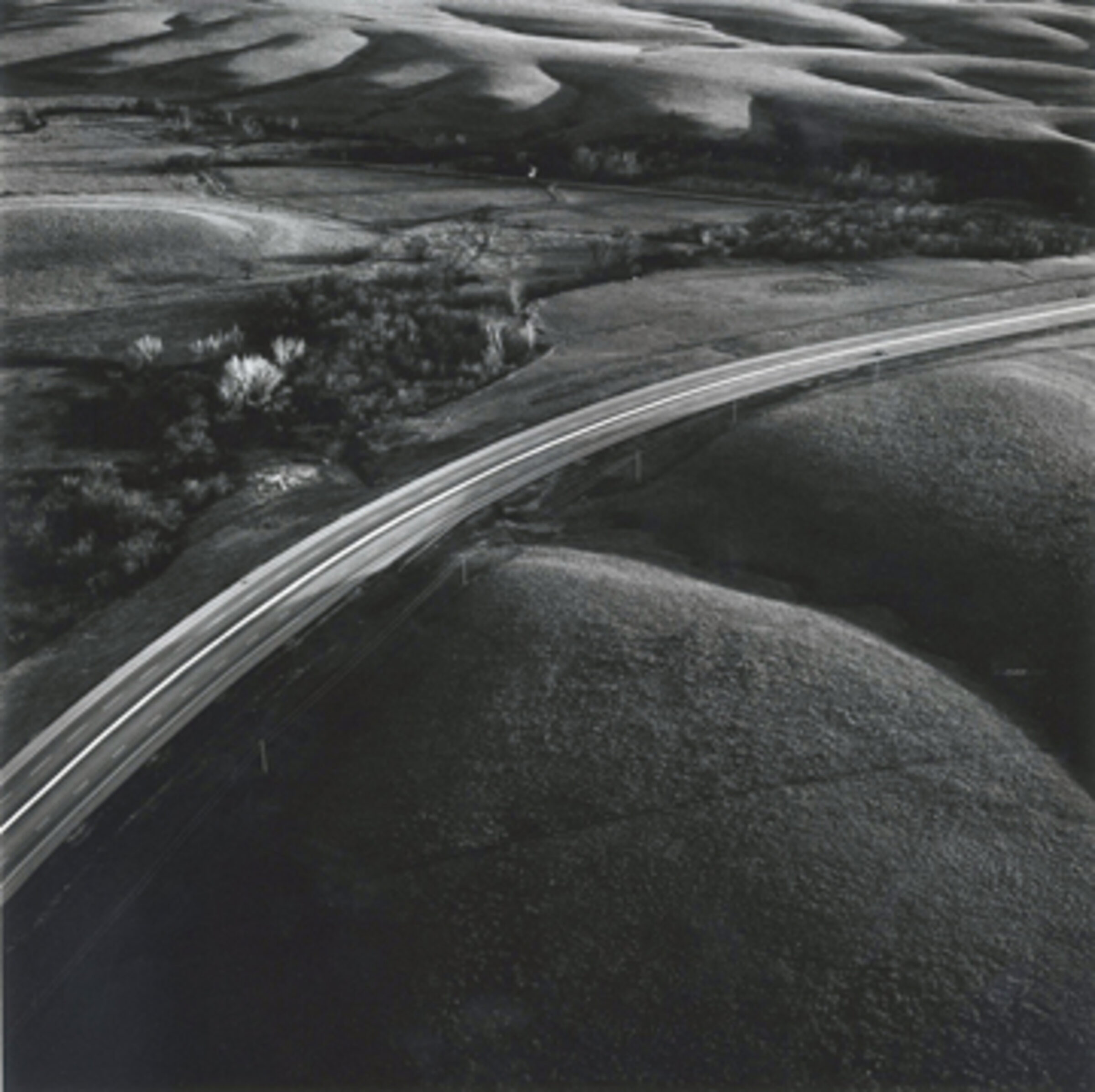 Aerial black and white photograph of a curved highway cutting through rolling prairie landscape, with agricultural fields visible in the distance and a cluster of trees and buildings nestled in a valley alongside the road.