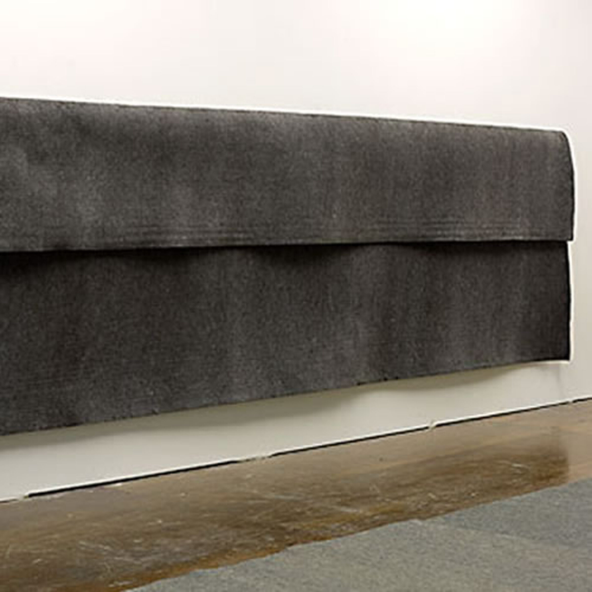 Large sheet of dark gray industrial felt suspended horizontally from a wall, with a pronounced fold creating a three-dimensional drape. The fabric hangs against a white wall above a wooden floor, emphasizing its weight and textural surface.