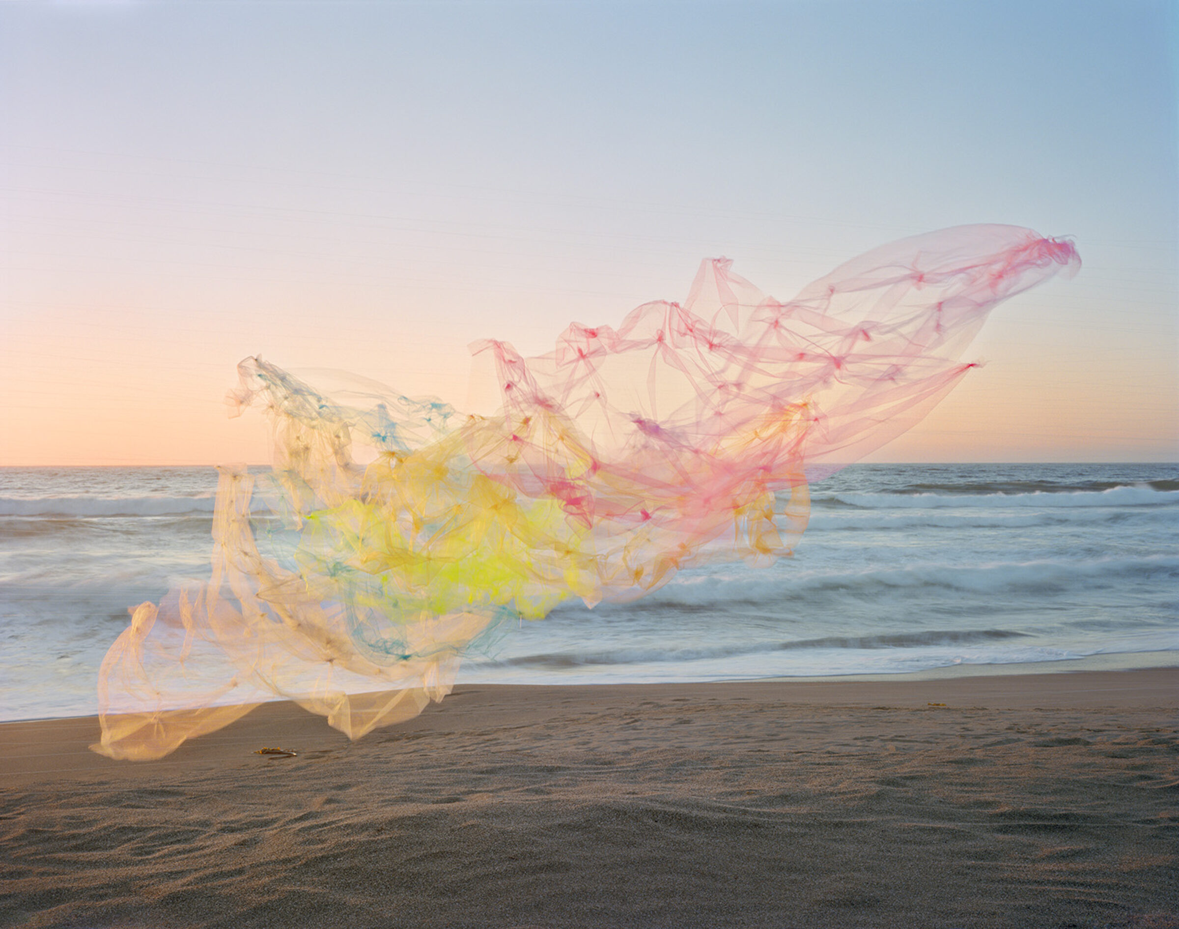 Billowing tulle fabric in soft gradations of pink, yellow, and blue floats across a beach at golden hour, with the ocean and horizon visible beyond. The translucent, flowing material creates an ethereal, sculptural form against the sand and seascape.