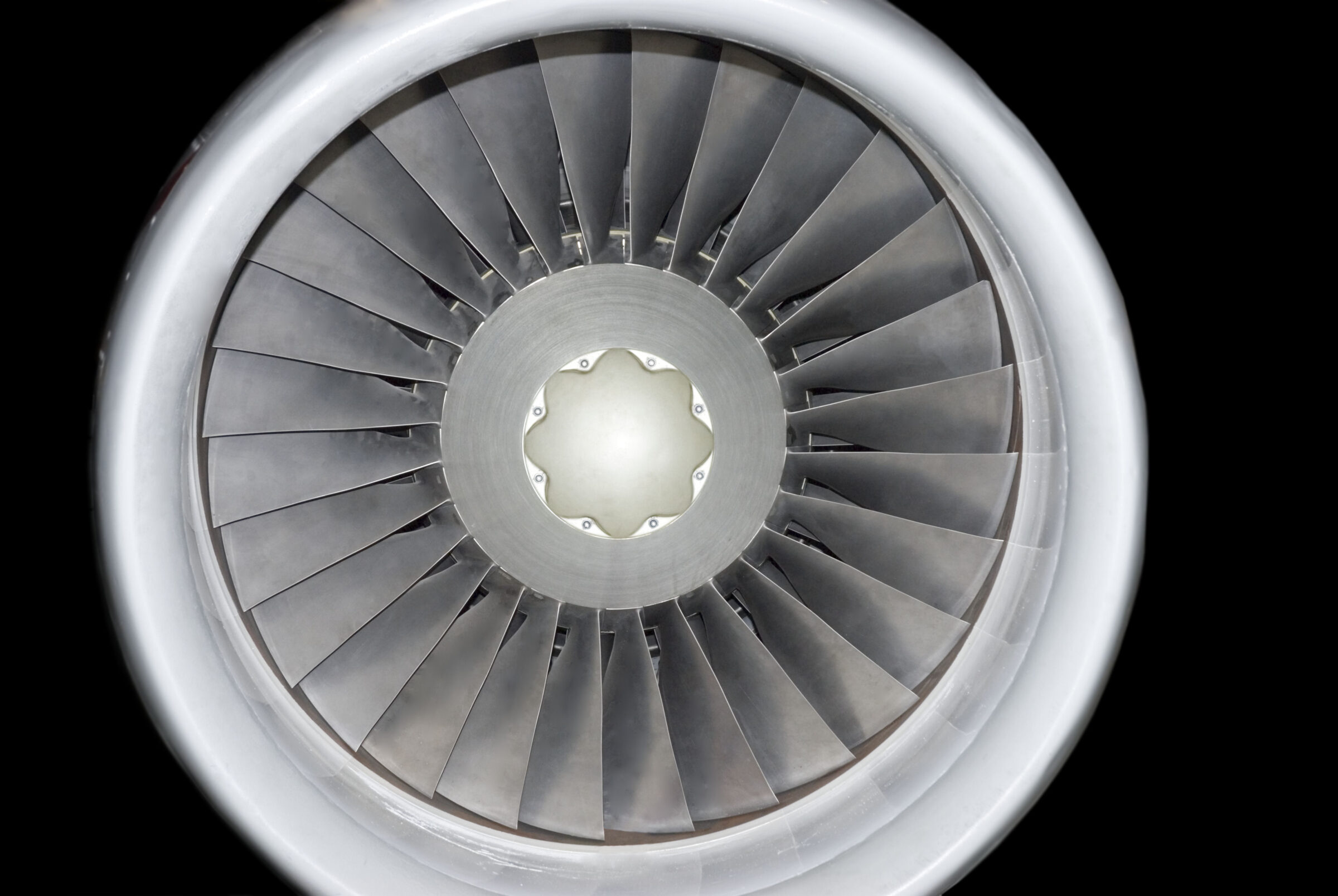 Front-facing view of a jet engine turbine with concentric metal blades radiating from a central hub, rendered in grayscale tones against a black background. The precise, symmetrical composition emphasizes the industrial geometry and reflective surfaces of the machinery.