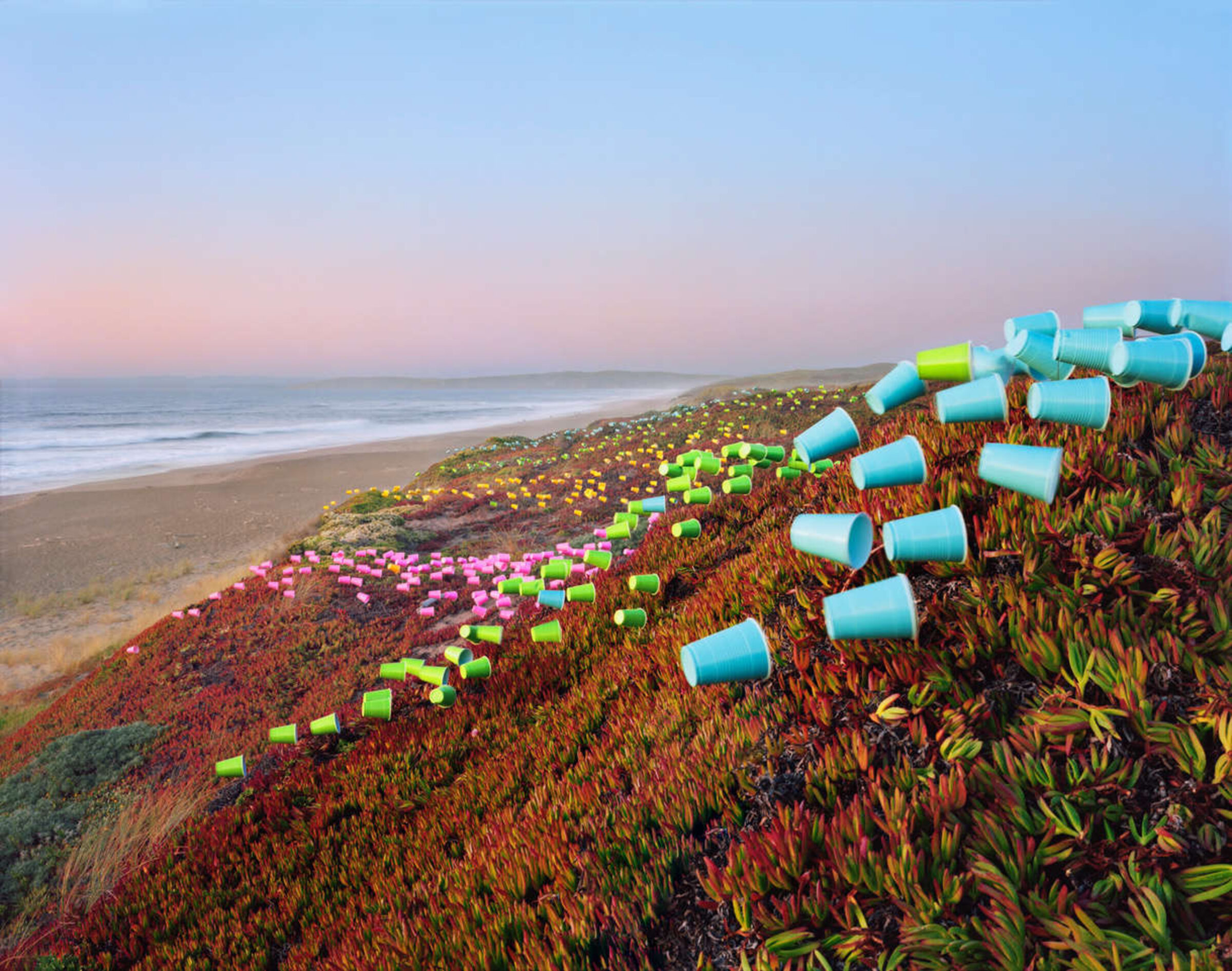 Colorful plastic cups arranged in geometric patterns across a coastal hillside covered in succulent vegetation, with a beach and ocean visible in the background at dusk.