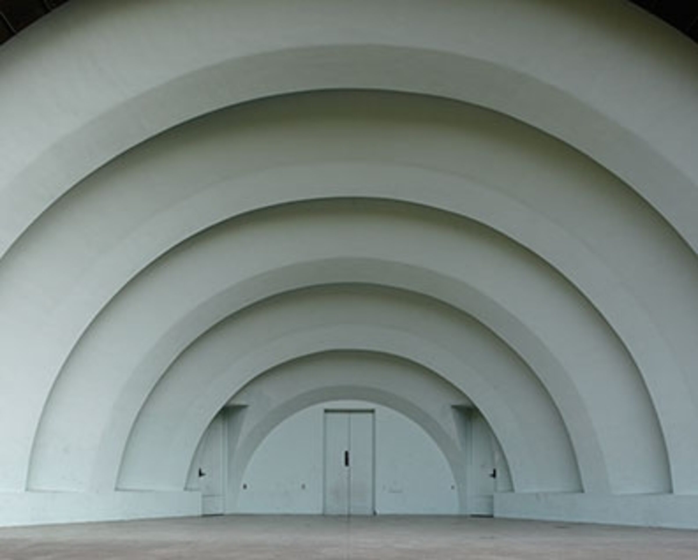 A series of concentric white arched structures recede toward a distant doorway in the center, creating a tunnel-like perspective. The geometric composition emphasizes depth and symmetry through repeated curved architectural forms against a minimal, monochromatic interior space.