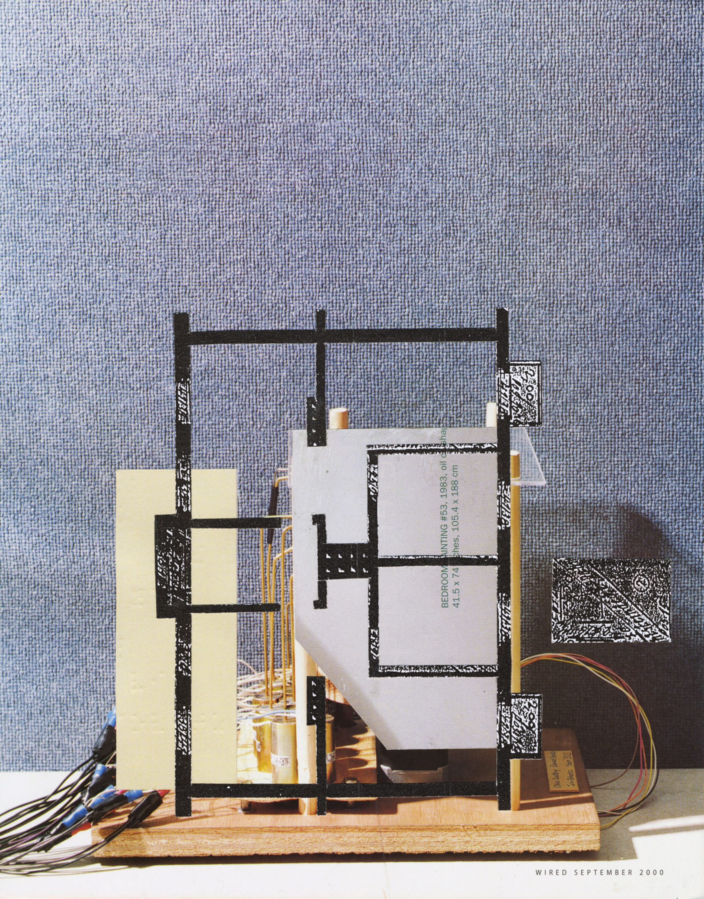 A three-dimensional collage construction mounted on a wooden base features black linear frameworks resembling architectural models, cream-colored paper planes, white rectangular forms with printed text, and wired electronic components in orange and red. Against a textured blue-gray backdrop, the assembled elements create a layered, abstract composition blending architectural, industrial, and mixed-media aesthetics.