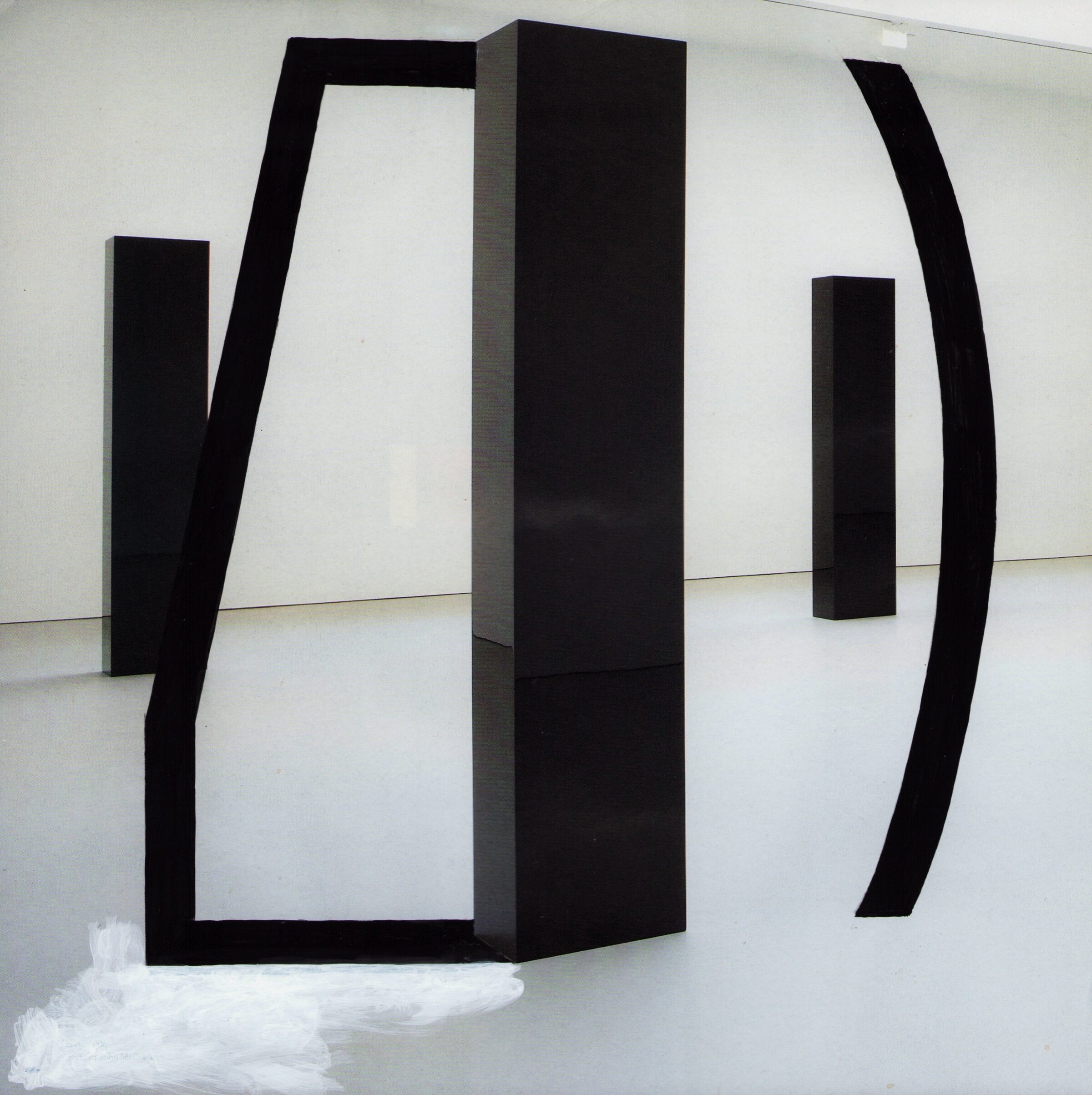 Black geometric forms constructed from stacked magazine pages stand in a gallery space with white walls and floor. The sculptural arrangement features angular and curved elements that create an open, architectural composition with negative spaces, accompanied by white material at the base.