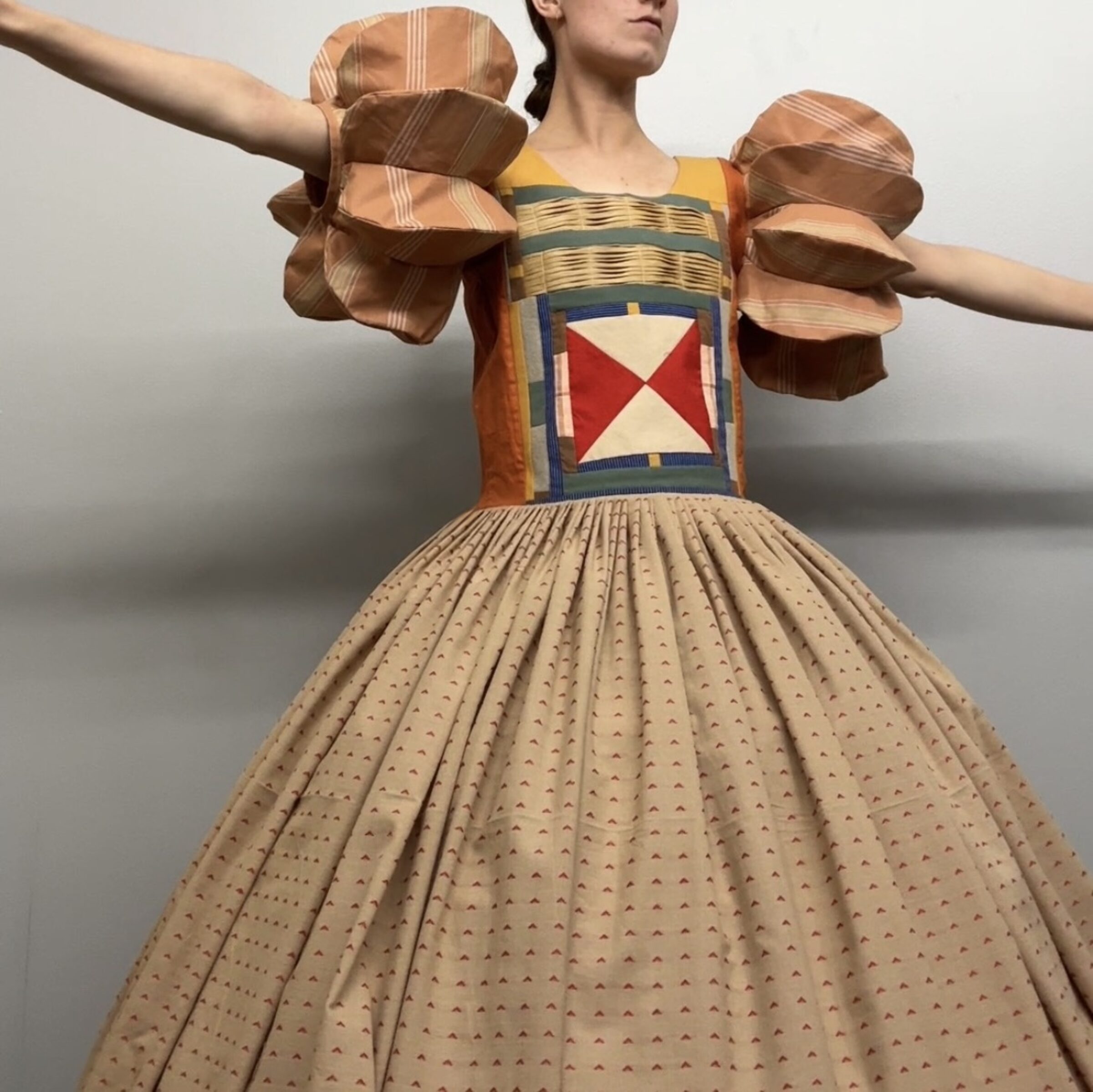 A figure in an elaborate costume stands with arms extended, wearing a voluminous pleated skirt in cream with tiny repeated patterns and a structured bodice featuring geometric patchwork in red, cream, navy, and gold tones. Large sculptural shoulder pieces in striped brown and tan fabric extend outward, creating a dramatic silhouette against a neutral background.