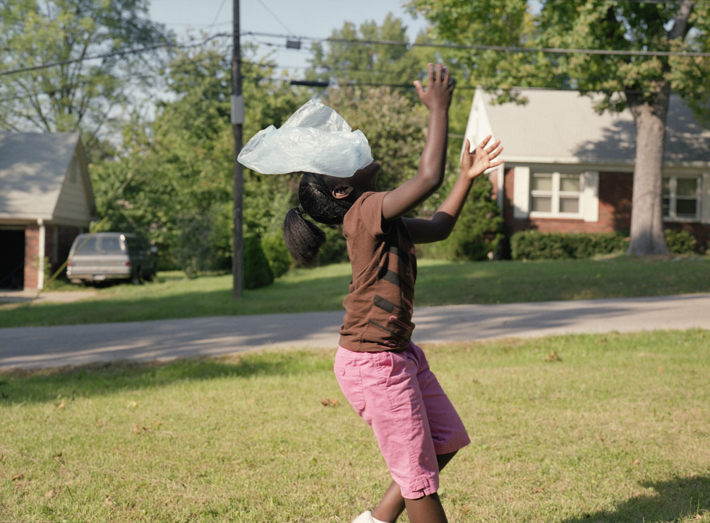 A figure stands in a residential street wearing a brown shirt and pink pants, with their head completely covered by a translucent white plastic bag. Their arms are raised expressively against a backdrop of tree-lined houses and suburban architecture.