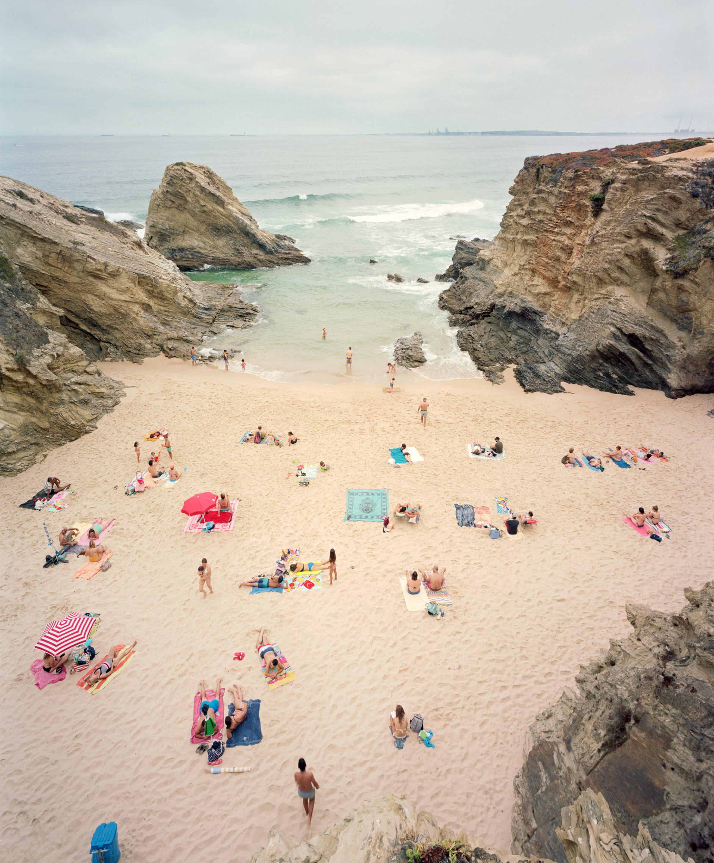 Aerial view of a sandy beach cove enclosed by dramatic rocky cliffs, with scattered beachgoers, colorful umbrellas, and towels dotting the sand. The turquoise and white waters beyond are studded with rocks, under an overcast sky.