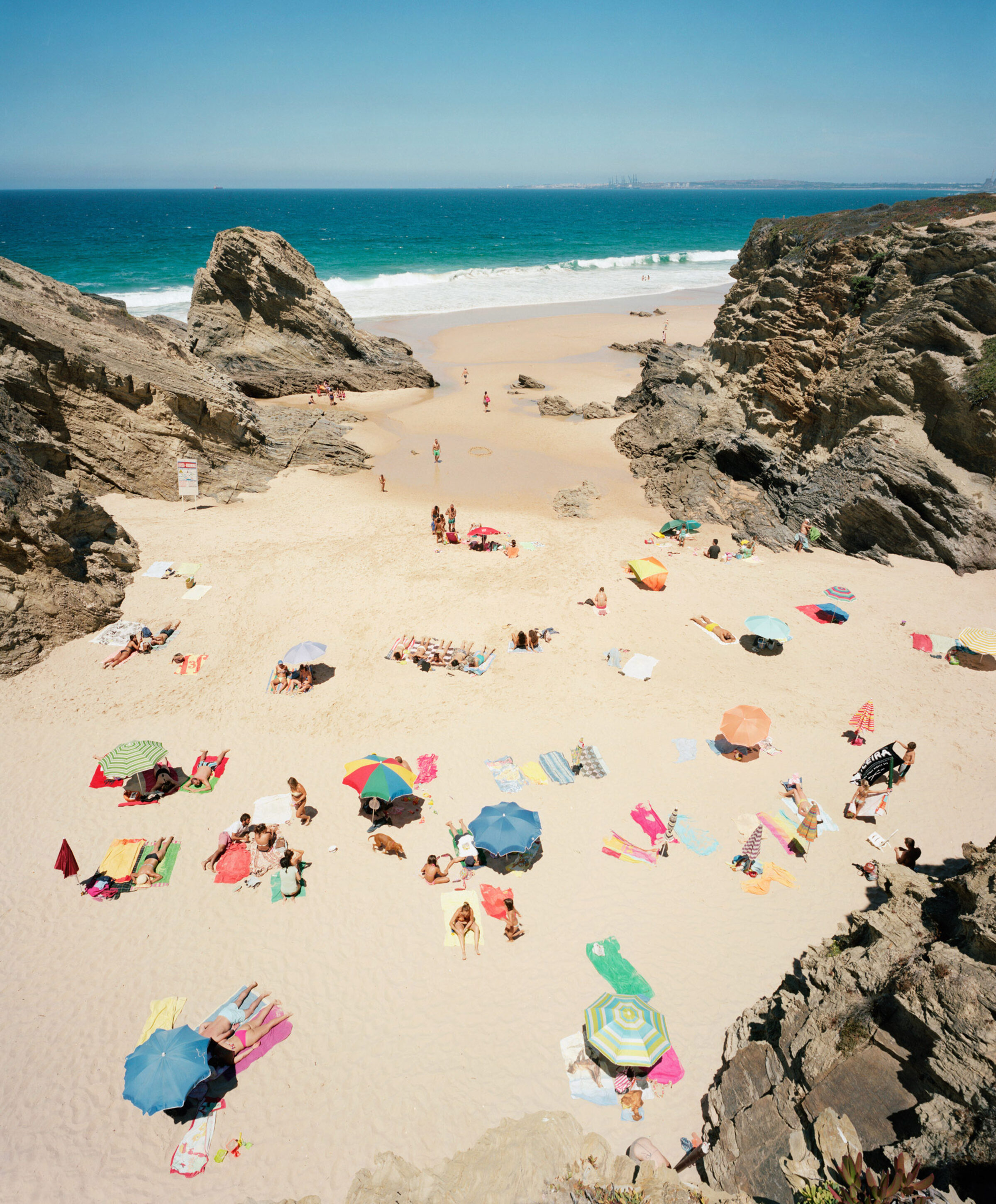 Aerial view of a secluded beach cove framed by dramatic rocky outcroppings, with numerous sunbathers scattered across the sand beneath a vibrant array of colorful beach umbrellas and towels in shades of pink, blue, yellow, and green. Turquoise waves break along the shoreline while the horizon extends to a distant coastline under clear blue sky.