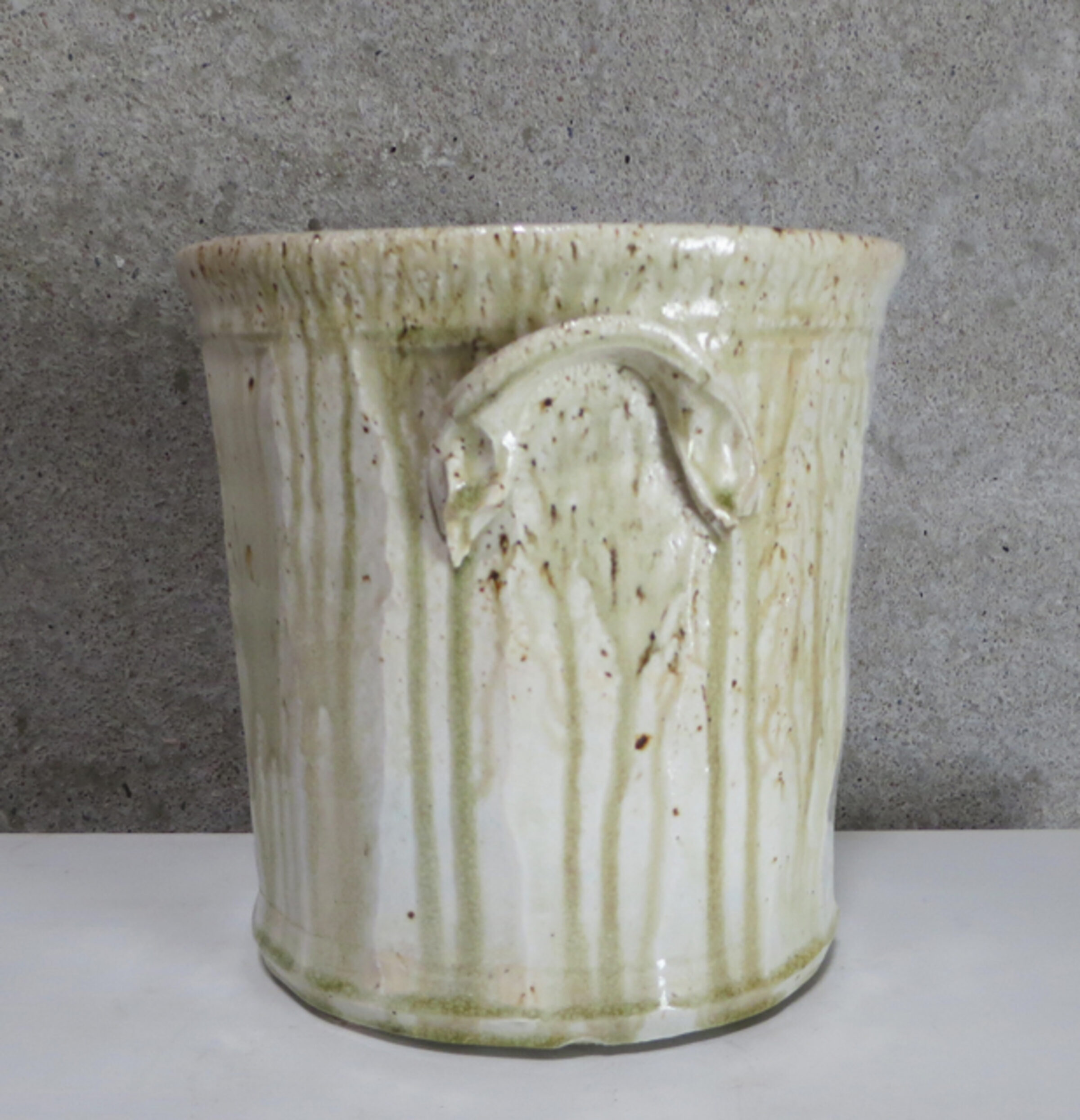 Cylindrical porcelain vessel with a cream-colored glaze featuring vertical striations and organic drip patterns, accented with brown speckles. The piece displays a subtle handle and exhibits the warm, natural variations characteristic of wood-fired ceramics.