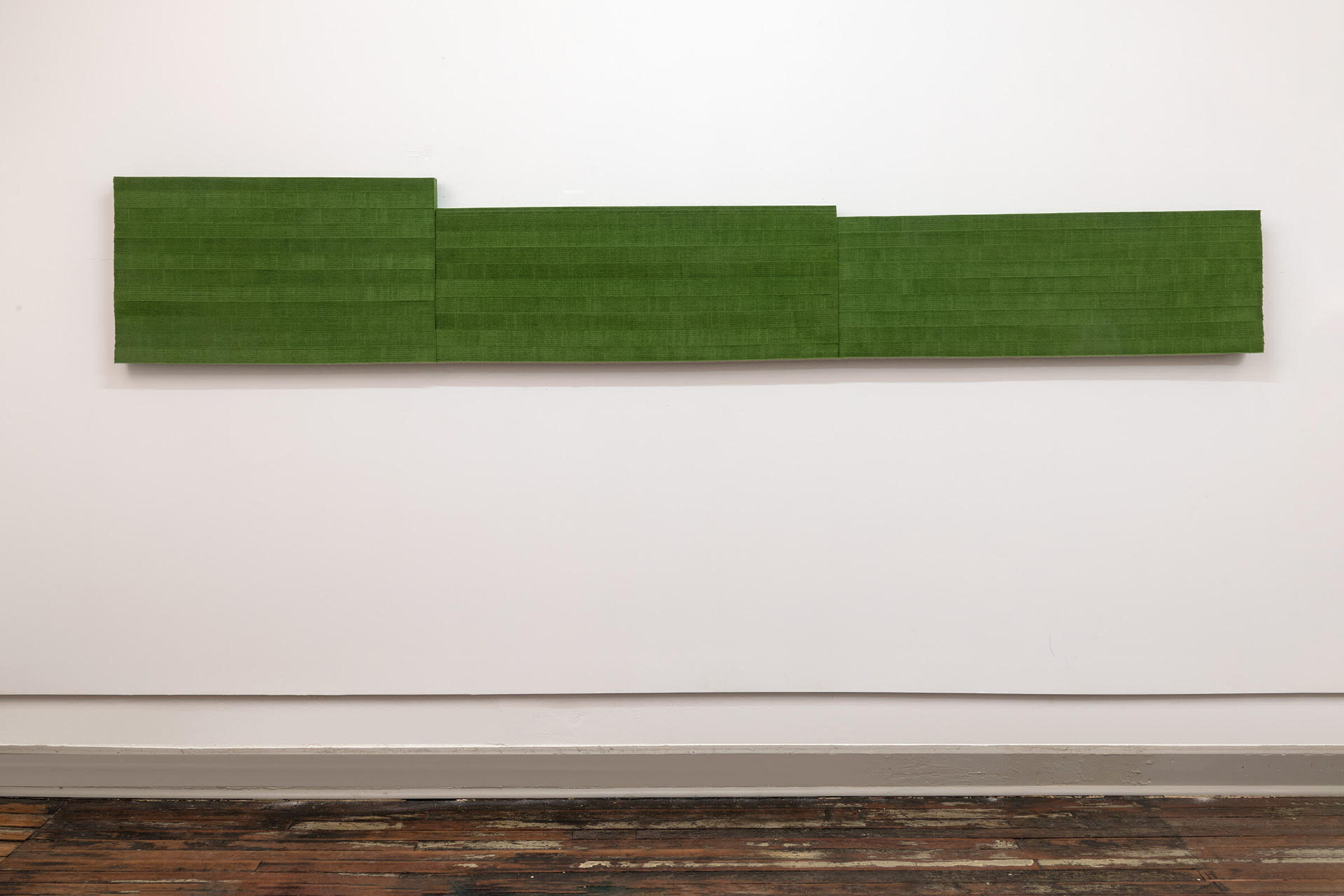 A horizontal green wool felt panel with stepped edges mounted on a white gallery wall above dark wooden flooring. The work consists of three rectangular sections of varying heights arranged in a staggered composition, creating a subtle architectural rhythm across the wall.