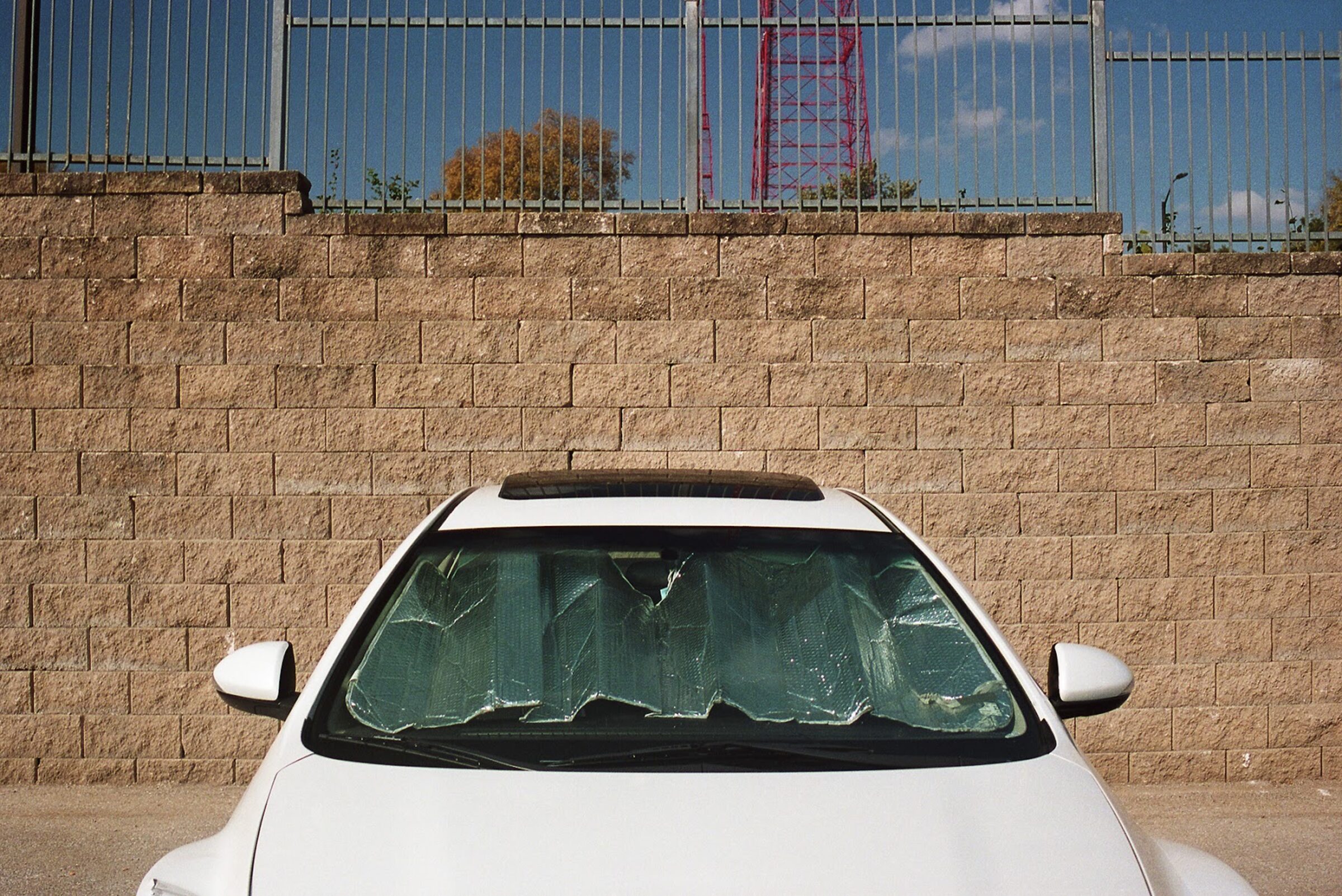 White sedan parked against a tan brick wall with a green sunshade covering its windshield, industrial metal fencing and red transmission tower visible in the background under a blue sky.
