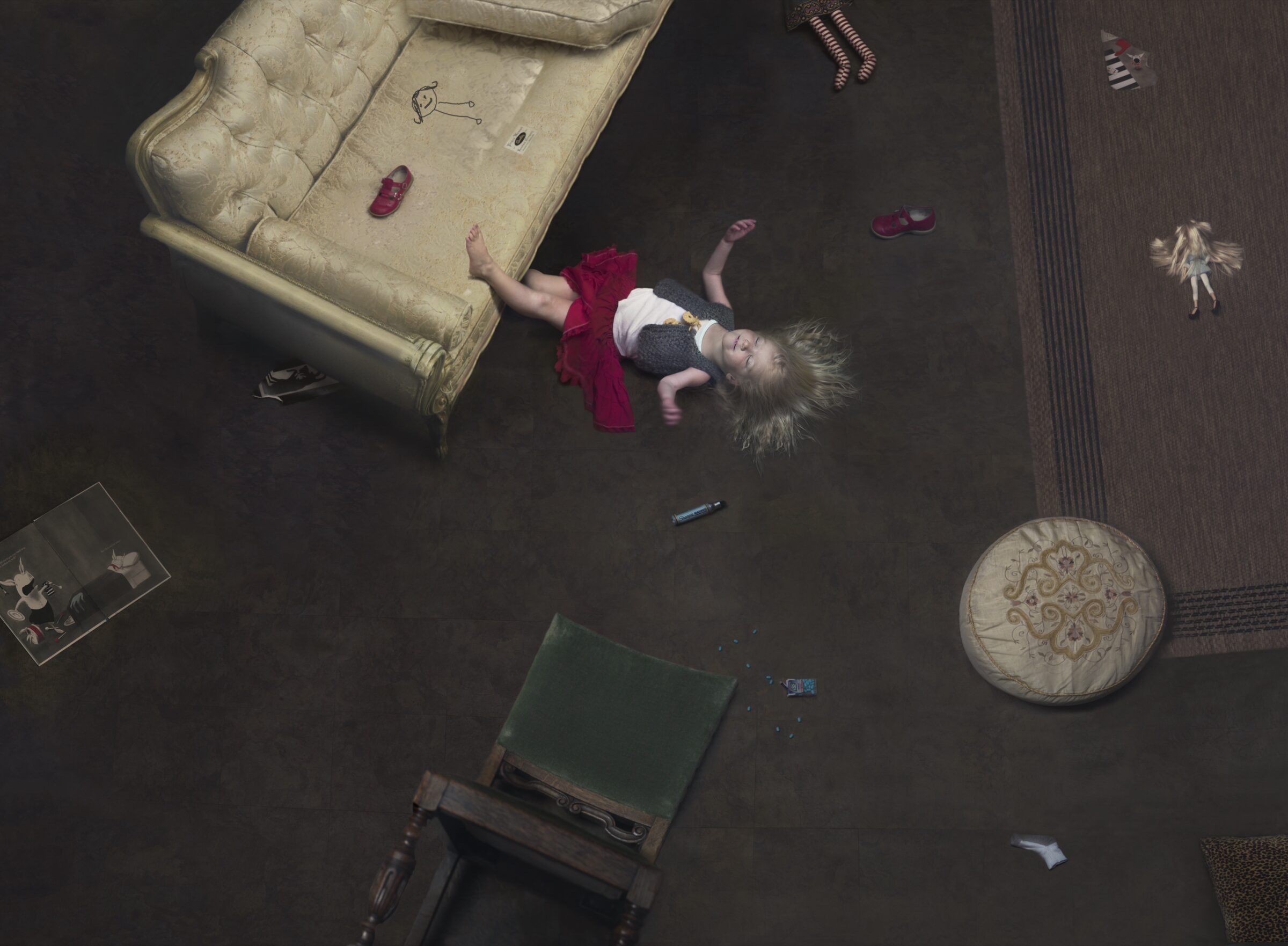Overhead view of a child lying on a dark floor surrounded by furniture and scattered objects, wearing a red shirt with arms raised. A cream-colored upholstered sofa, round ottoman, green chair, and various items including shoes, drawings, and small objects are arranged around the figure.