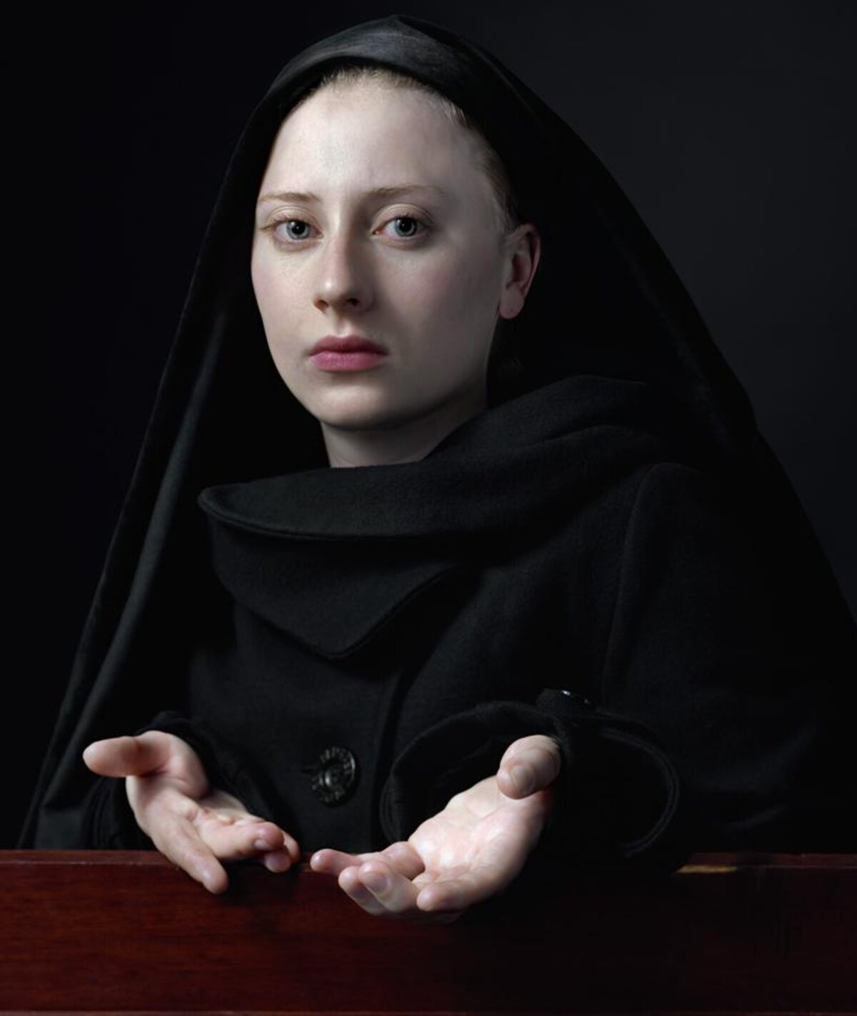 A pale-skinned figure wearing a black hooded cloak gazes directly forward with a neutral expression against a dark background. Their hands are positioned open and gesturing toward the viewer across a rich wooden surface, creating an intimate yet enigmatic portrait composition.
