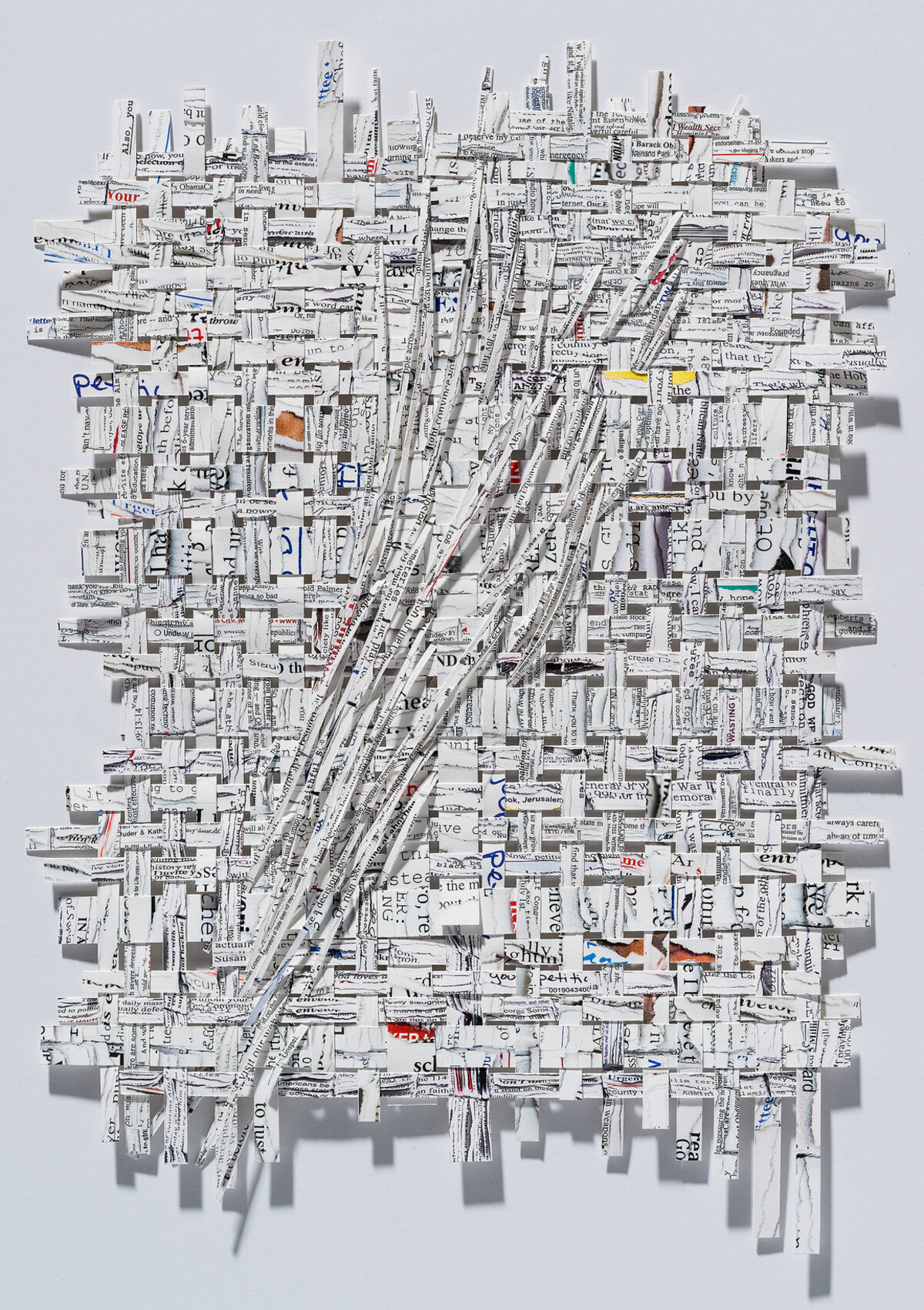 A three-dimensional collage of shredded and layered printed text and newspaper fragments arranged on a white wall, creating a dense, textured composition with diagonal striations and scattered edges. Occasional pops of color—including yellow, orange, red, and blue accents—punctuate the predominantly white and gray tones.