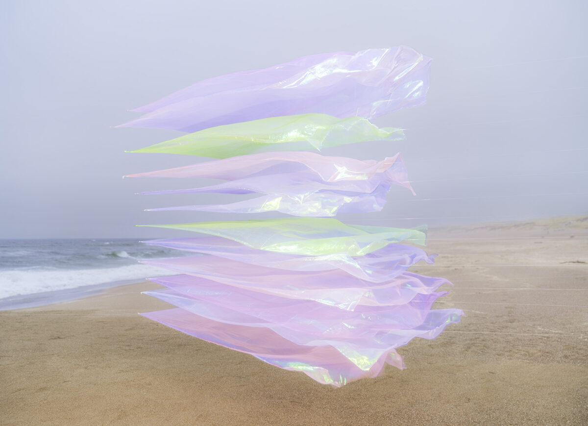 Translucent iridescent fabric strips in shades of lavender, pale green, and pink are layered and suspended vertically above sand, with an ocean and shoreline visible in the soft-focused background.