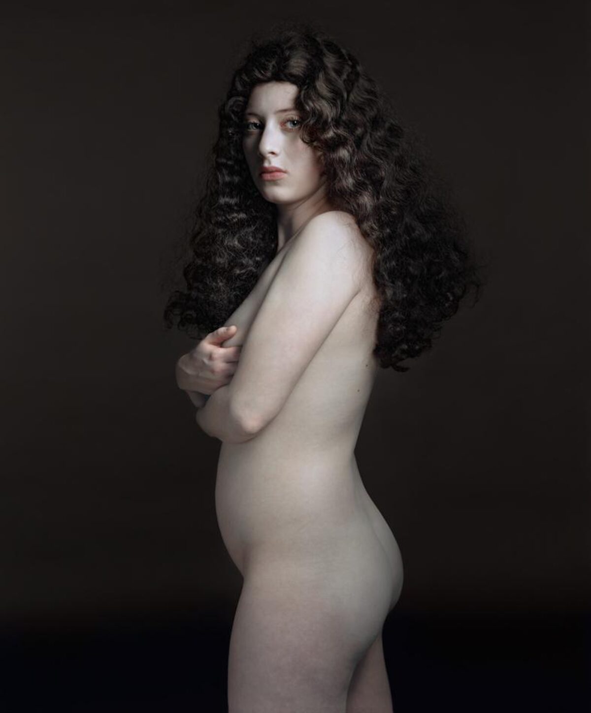 Portrait of a woman with long, voluminous dark curly hair shown in profile against a dark neutral background. She is posed with her body turned away and arm crossed, rendered in pale tones that create strong contrast with the shadowed backdrop.
