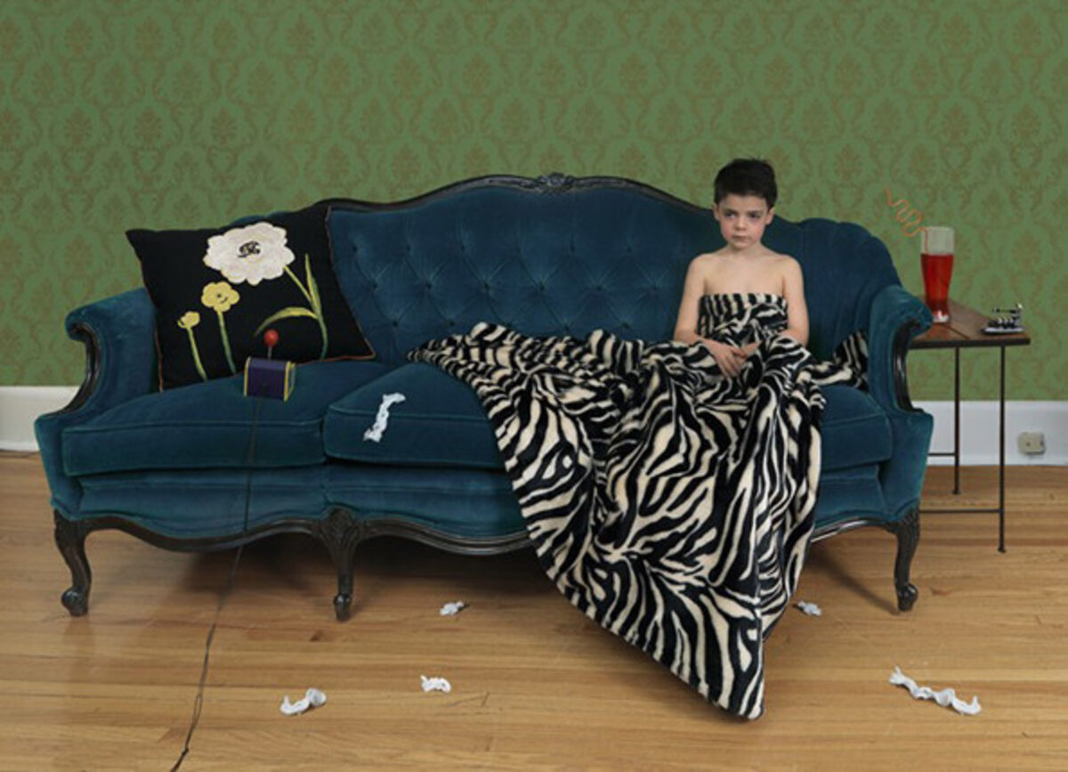 A person in a black strapless top reclines on a teal upholstered sofa draped with a zebra-striped blanket, against a green damask wallpapered wall. Scattered tissues and objects lie on the wooden floor, while a side table with a glass holds personal items nearby, creating a scene of domestic illness or malaise.