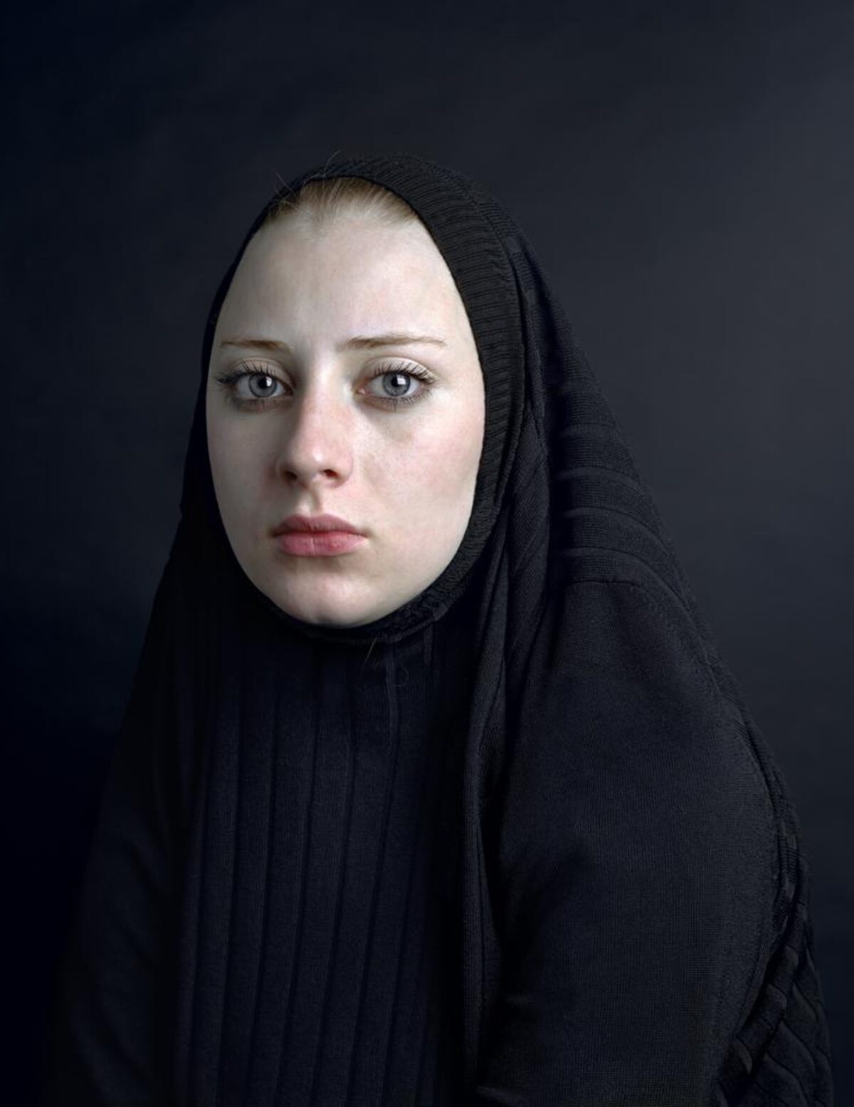 Portrait of a pale-skinned woman with blue eyes gazing directly at the camera, her head covered with a black textured fabric draped over her shoulders against a dark gray background. The lighting is stark and frontal, emphasizing the contrast between her fair complexion and the dark surrounding fabric.