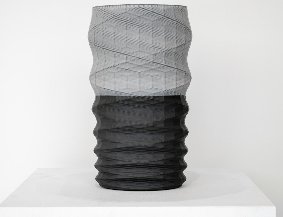 A tall cylindrical vessel with a twisted, ribbed surface sits on a white platform. The upper half features a translucent gray mesh-like texture, while the lower half is rendered in solid black with horizontal ridging, creating a striking color and material contrast.