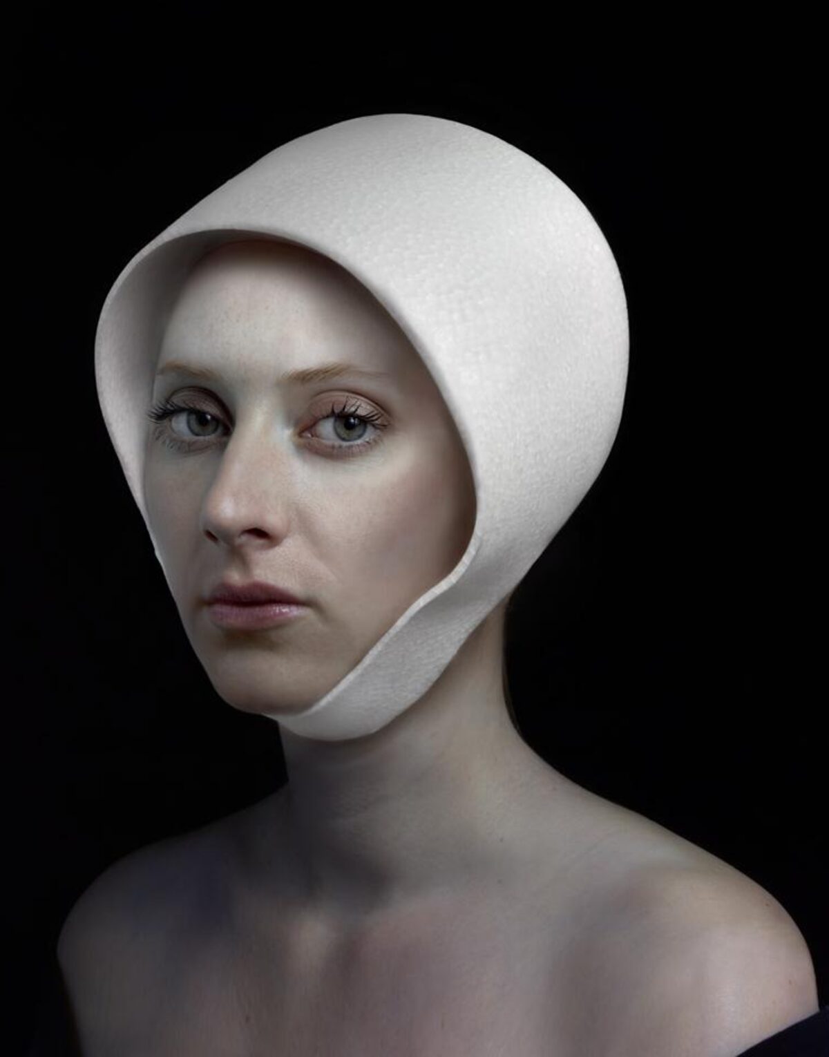 Portrait of a pale-skinned woman with dark eyes and lips against a black background, her head wrapped in a white fabric hood that frames her face. The minimalist composition emphasizes the subject's contemplative expression and the stark contrast between her skin tone and the pristine white textile.