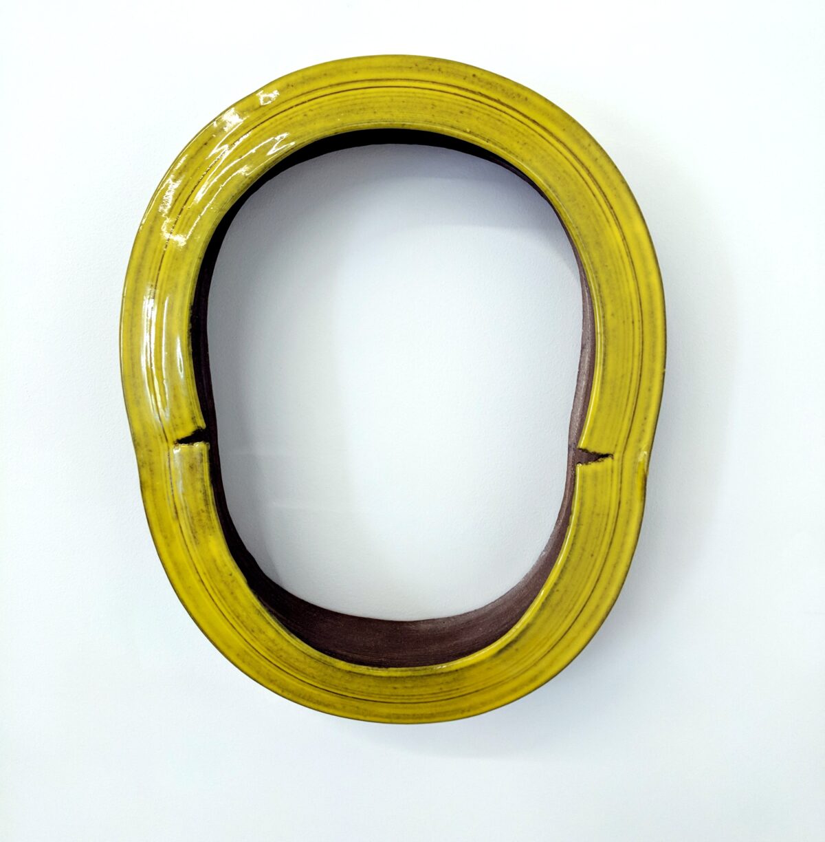 A vibrant yellow-green glazed ceramic ring form with an oval opening, set against a white background. The glossy surface contrasts with the darker interior, and subtle linear textures run across the frame's width.