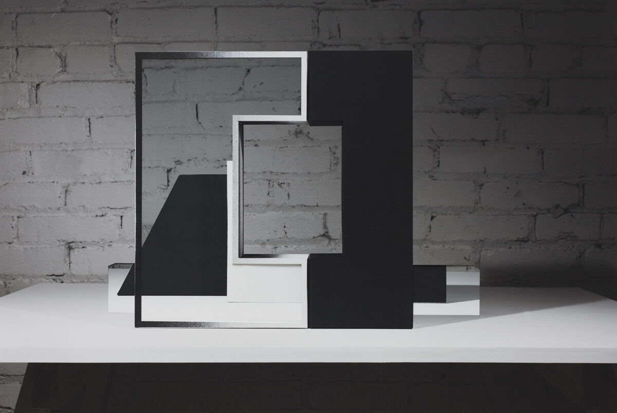 Abstract geometric composition of layered rectangular frames in black and white, creating a three-dimensional architectural form against a weathered brick wall. The overlapping planes and frames cast shadows on a white surface, establishing depth through contrasting solid and void spaces.
