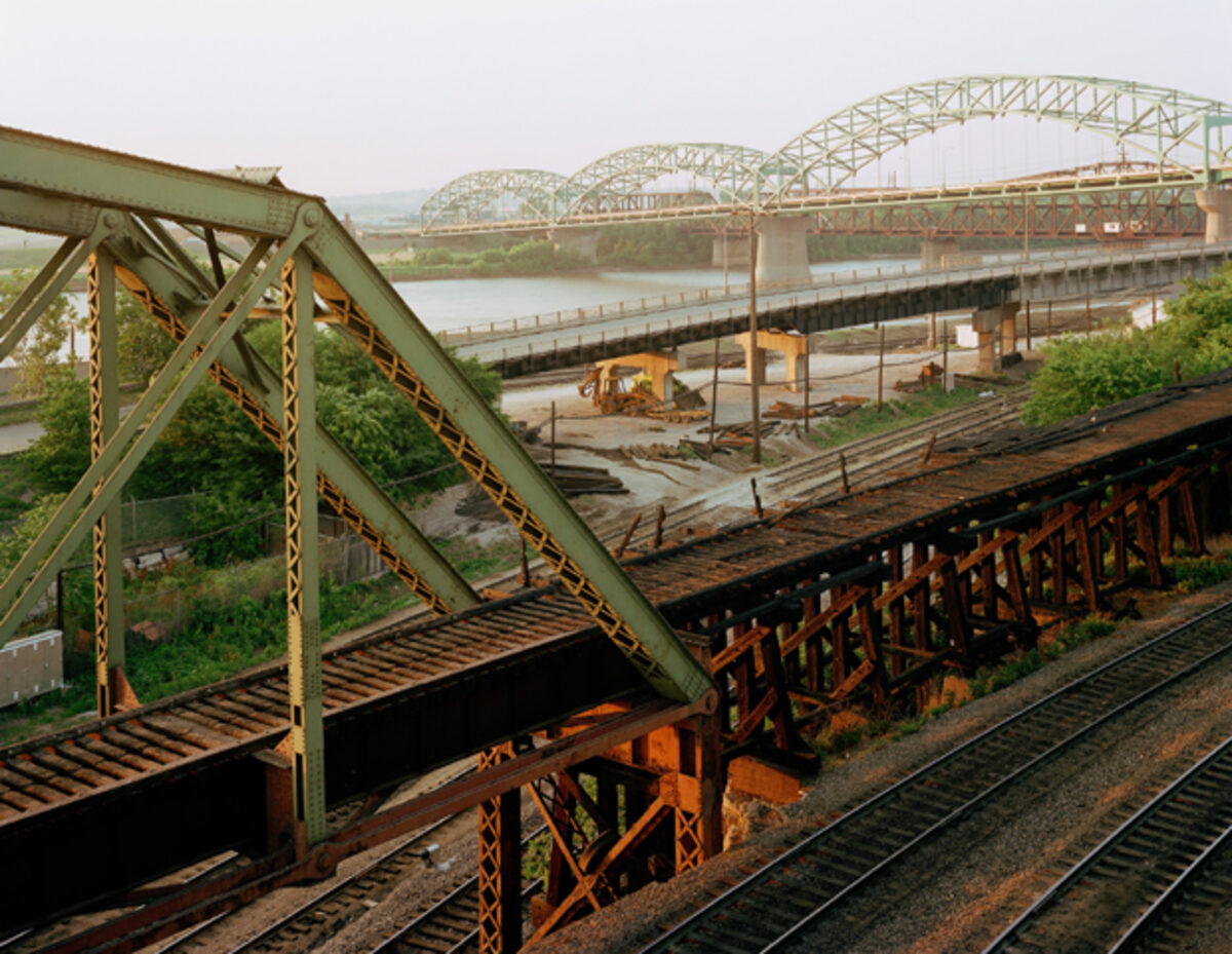 Industrial railroad bridge infrastructure in the foreground with rusted steel trusses and support beams, extending toward a modern arched bridge crossing a wide river in the distance. The composition layers deteriorating metalwork, active rail yards with equipment and structures, and waterway landscape under overcast sky, rendered in muted greens, browns, and grays.