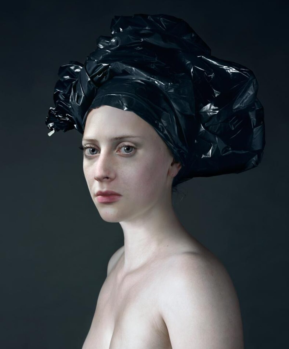Portrait of a pale-skinned person with a neutral expression, shown in three-quarter profile against a dark gray background. Their head is wrapped in crumpled black plastic material that extends upward and outward, creating an elaborate sculptural headdress. The figure's shoulders are bare, emphasizing the stark contrast between skin tone and the glossy black material.