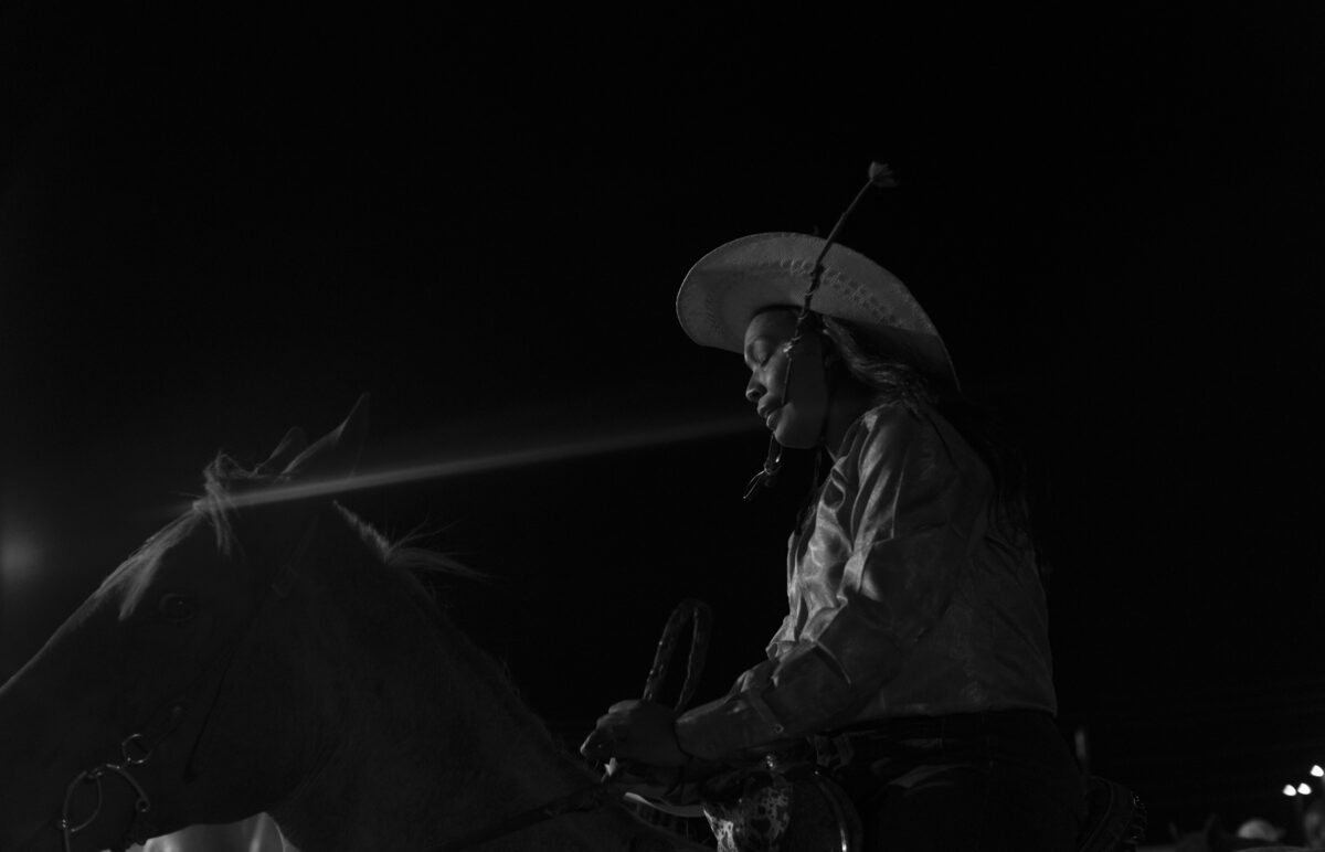 Black and white photograph of a figure wearing a wide-brimmed hat and long coat, seated on a horse against a dark background. The composition is rendered in high contrast, with dramatic lighting that emphasizes the silhouettes of both rider and horse.