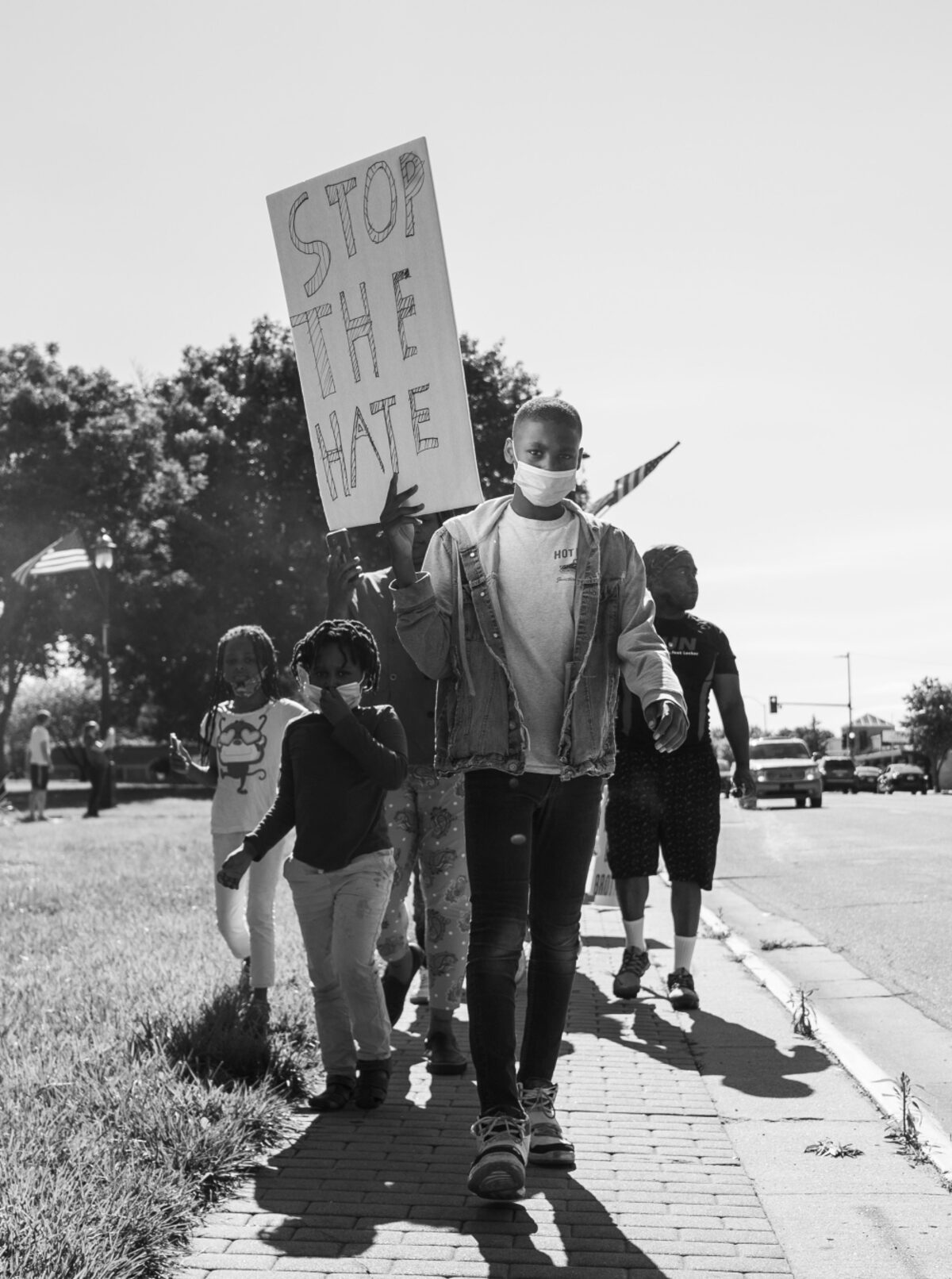 Black and white photograph of a group of people walking on a sidewalk, led by a young person wearing a face mask and holding a large sign reading "STOP THE HATE." Additional protesters, including children, walk alongside. Trees and parked cars are visible in the background.