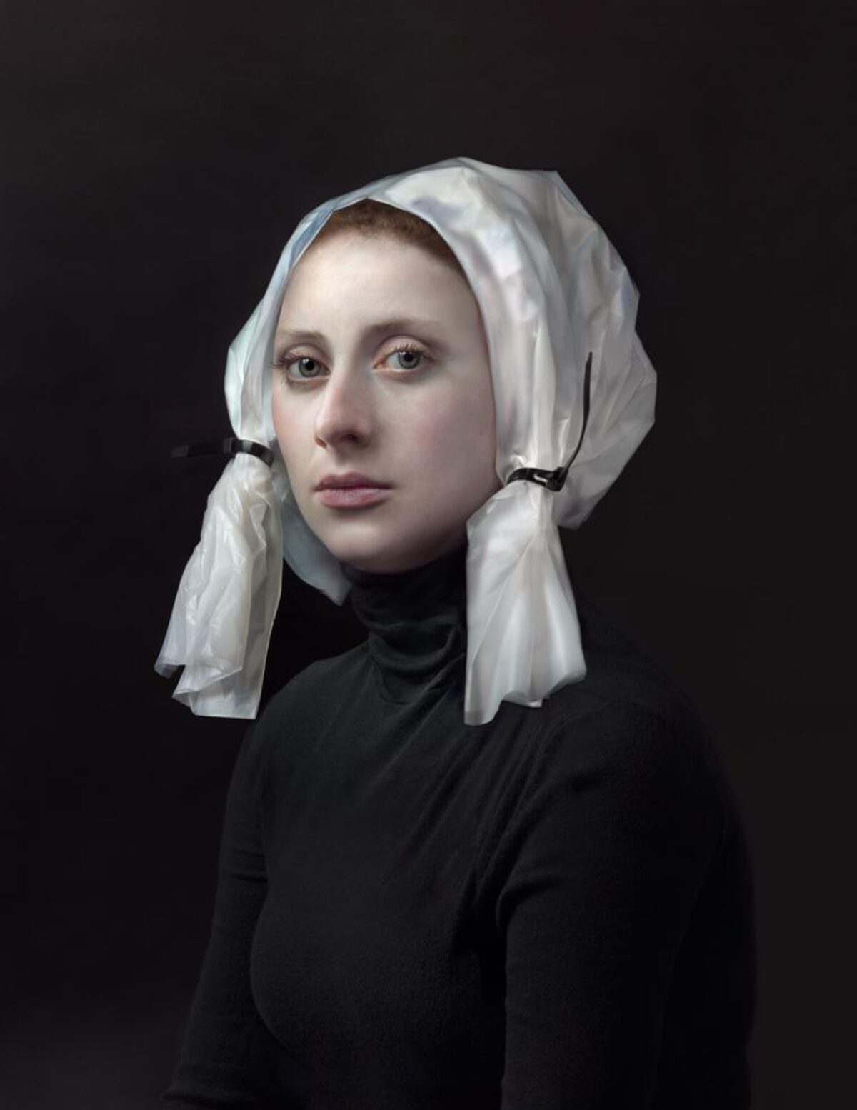 Portrait of a woman against a dark background, wearing a black turtleneck and a white linen head covering tied beneath the chin. Her gaze is directed slightly away from the camera, with a composed, contemplative expression.